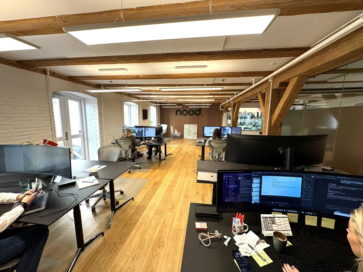 Man ser tre mennesker siddende ved arbejdsborde med computere. Der er flere arbejdsborde og de er alle besat af folk. På