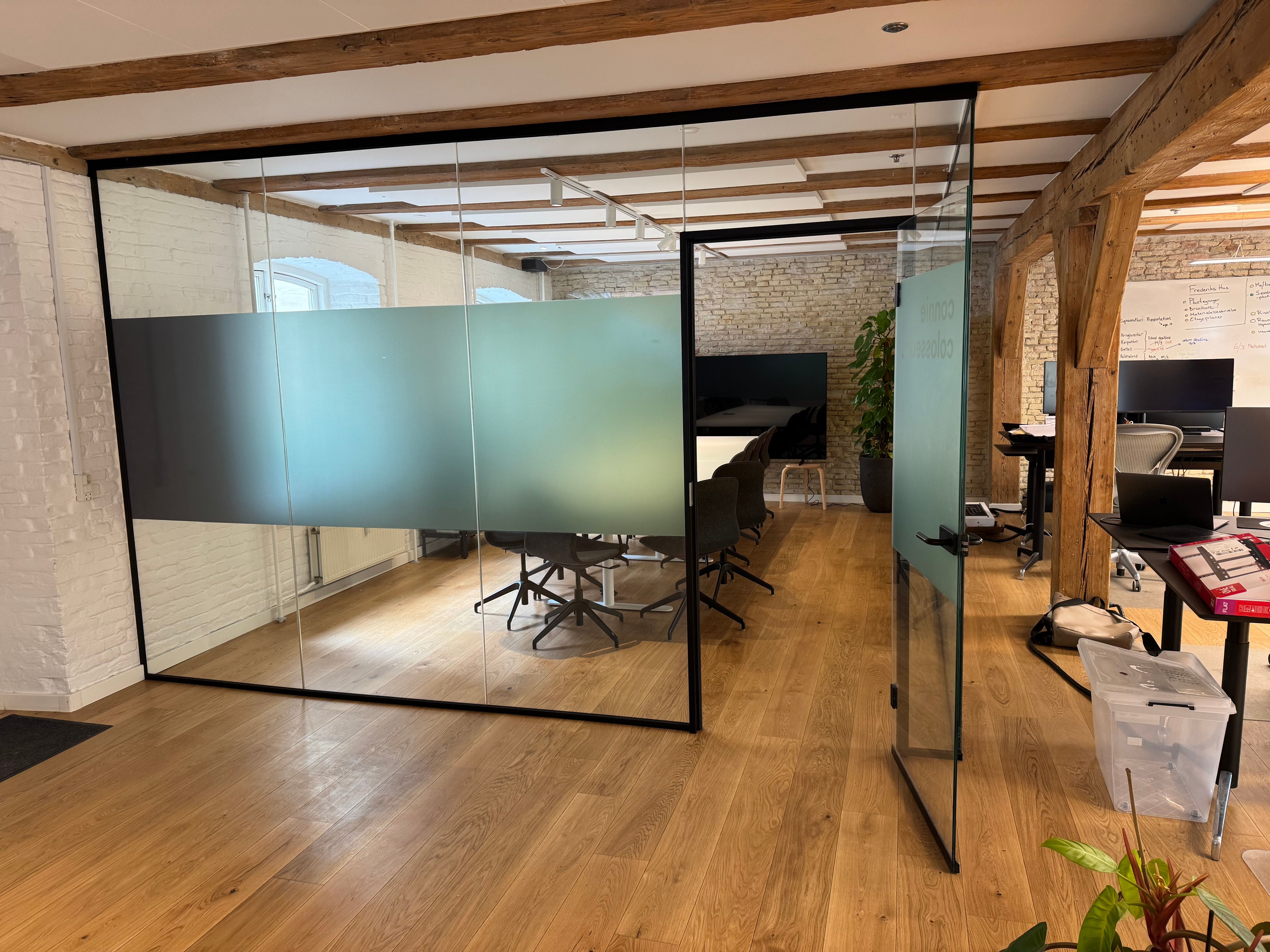 En moderne kontor med højt loft og åbent layout. Glasvægge og bord og stole. Et bord med en computer og en lampe. En plante