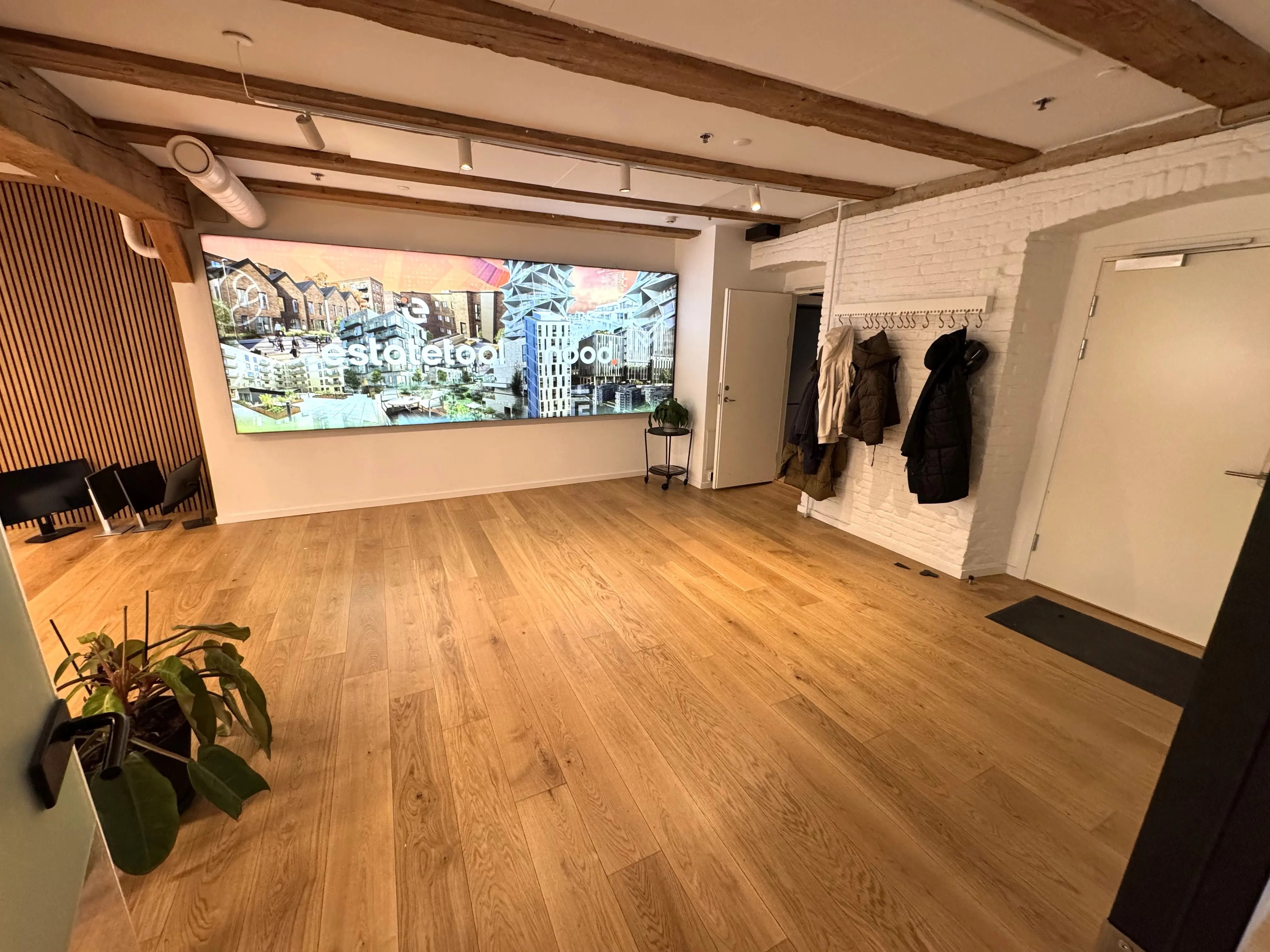 En stor stue med trægulv og lysekroner, en stor tv-skærm med en byudsigt, en hængeklædeskab med tøj og en plante på gulvet