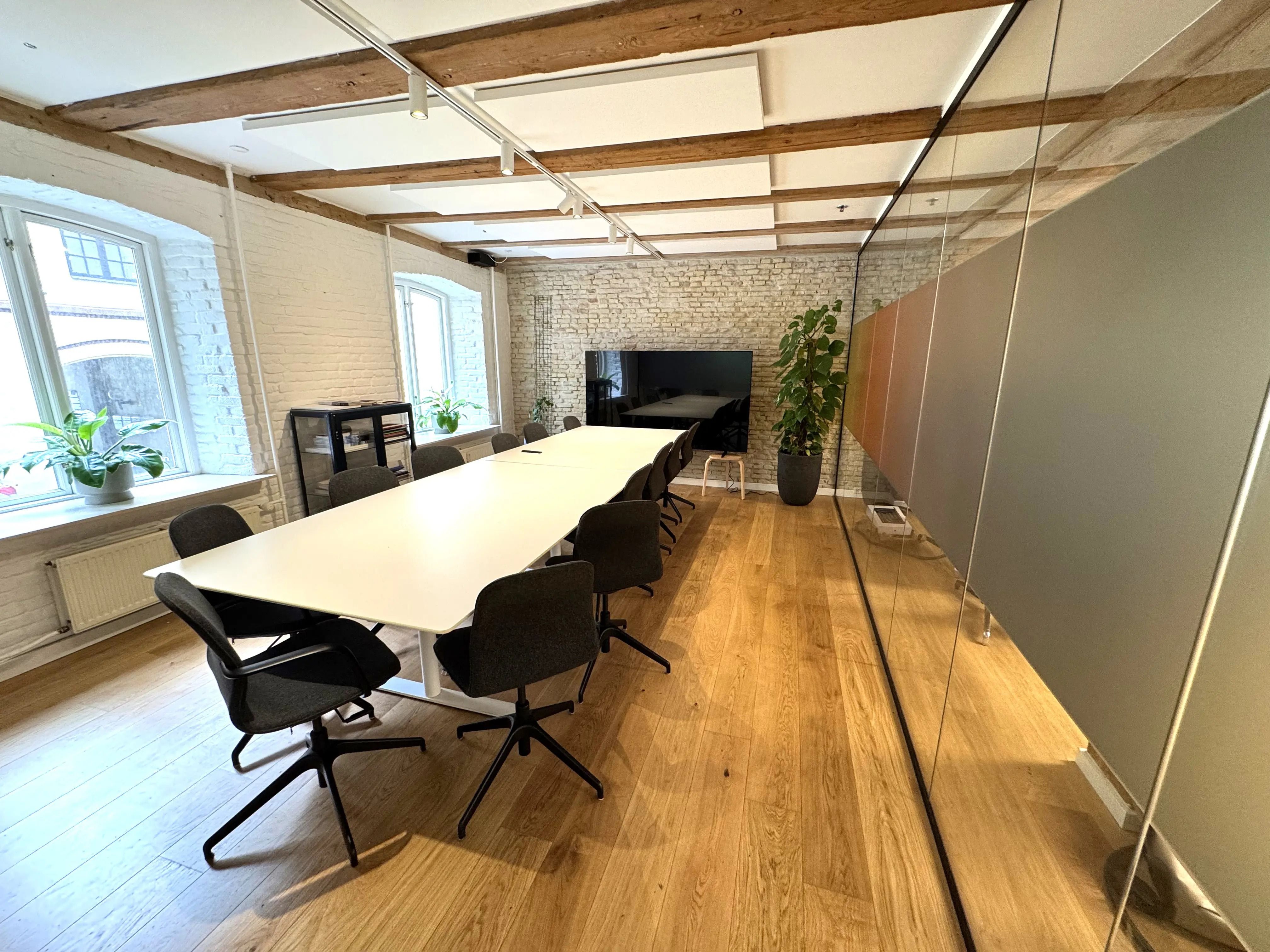 Et stort mødelokale med højt loft, hvide vægge og møbler med høj kvalitet samt flere plante og en stor tv-skærm