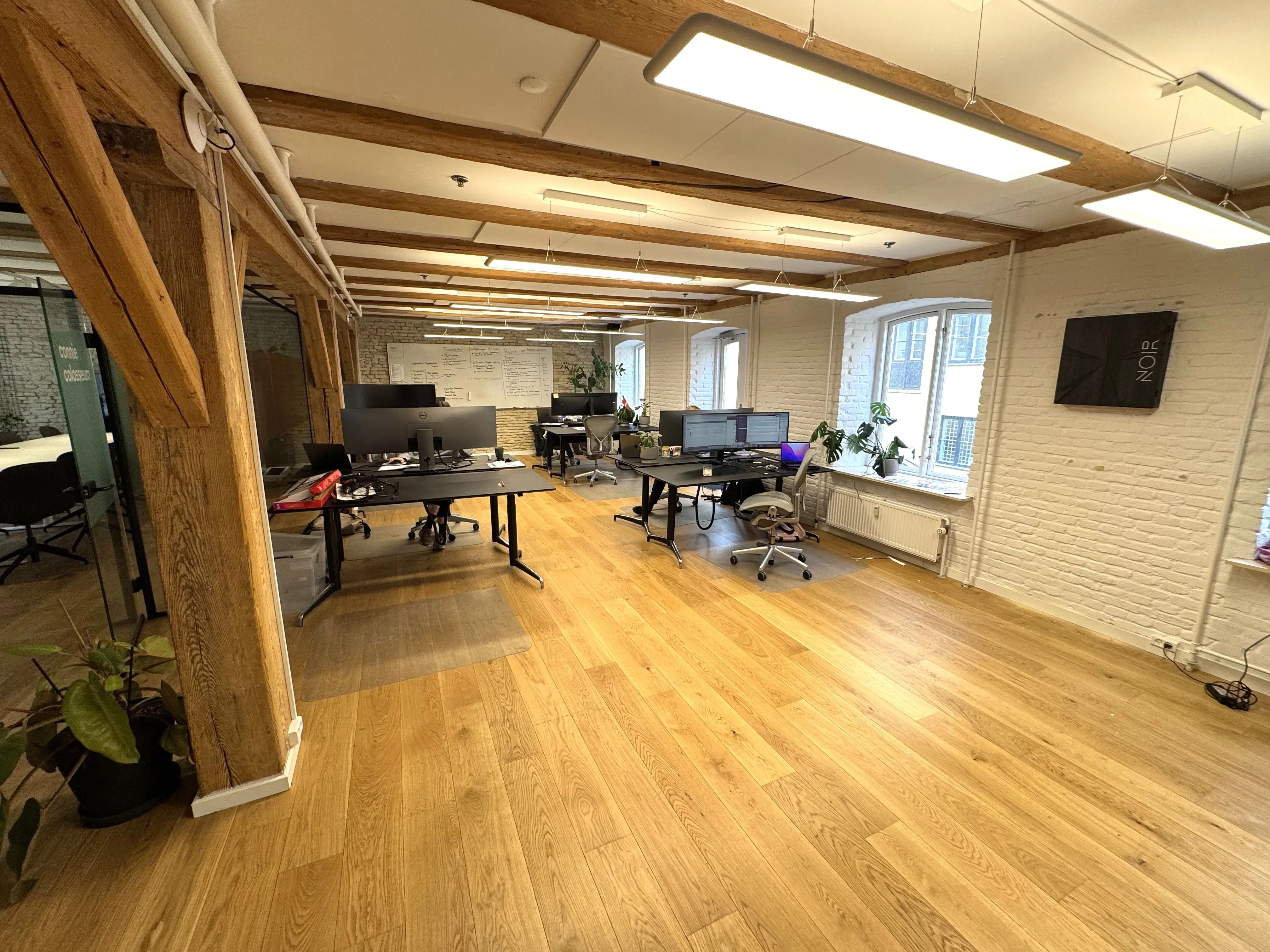 Ledig kontor med computere og skriveborde. Trægulve, hvide vægge og lyse loftsbælge. Plakat med "NORE" og plante på