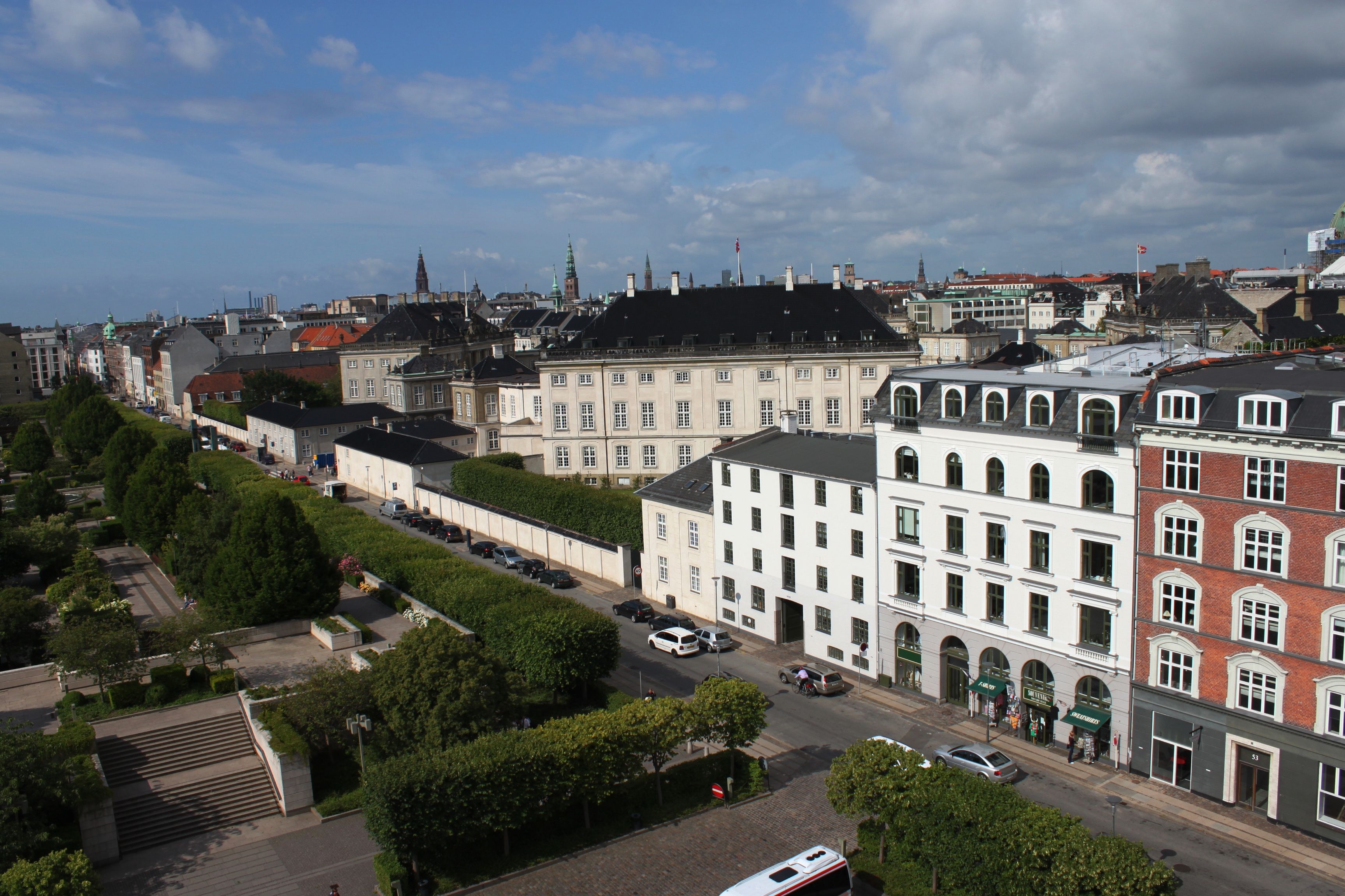 Flere høje bygninger med parker og biler parkeret langs gaden med en bus og personer på siden