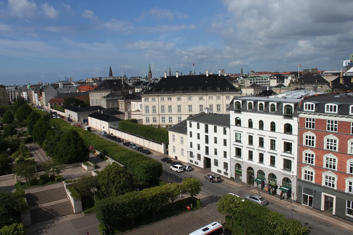 Flere høje bygninger med parker og biler parkeret langs gaden med en bus og personer på siden