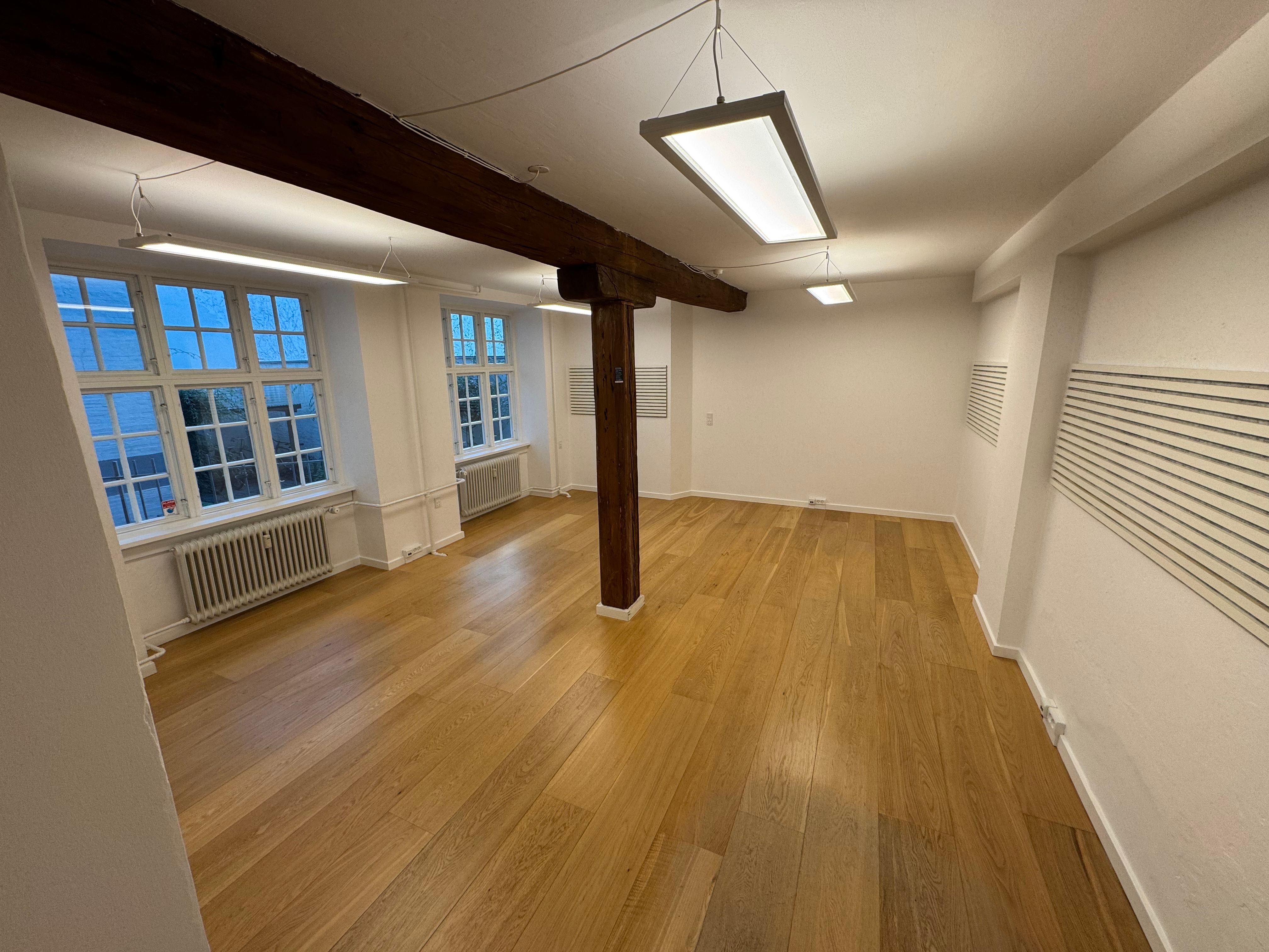 Kontor med hårdt trægulv, store vinduer, lyse og lyse gulvlampe og lyse hvide vægge.