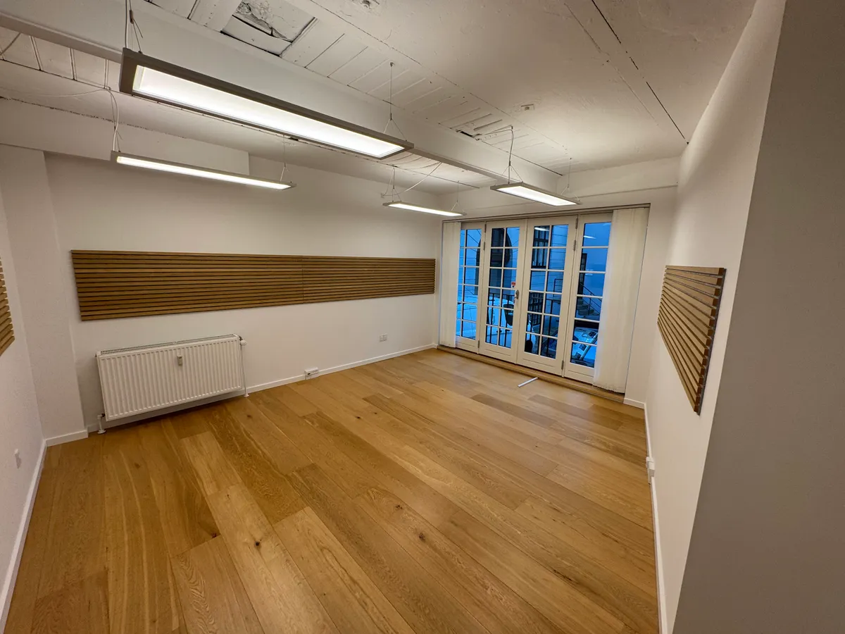 Ledig kontorrum med lyse loftslampe, hvide vægge, lyse trægulve og store glasdøre. Der er et hvidt radiator og to træpaneler
