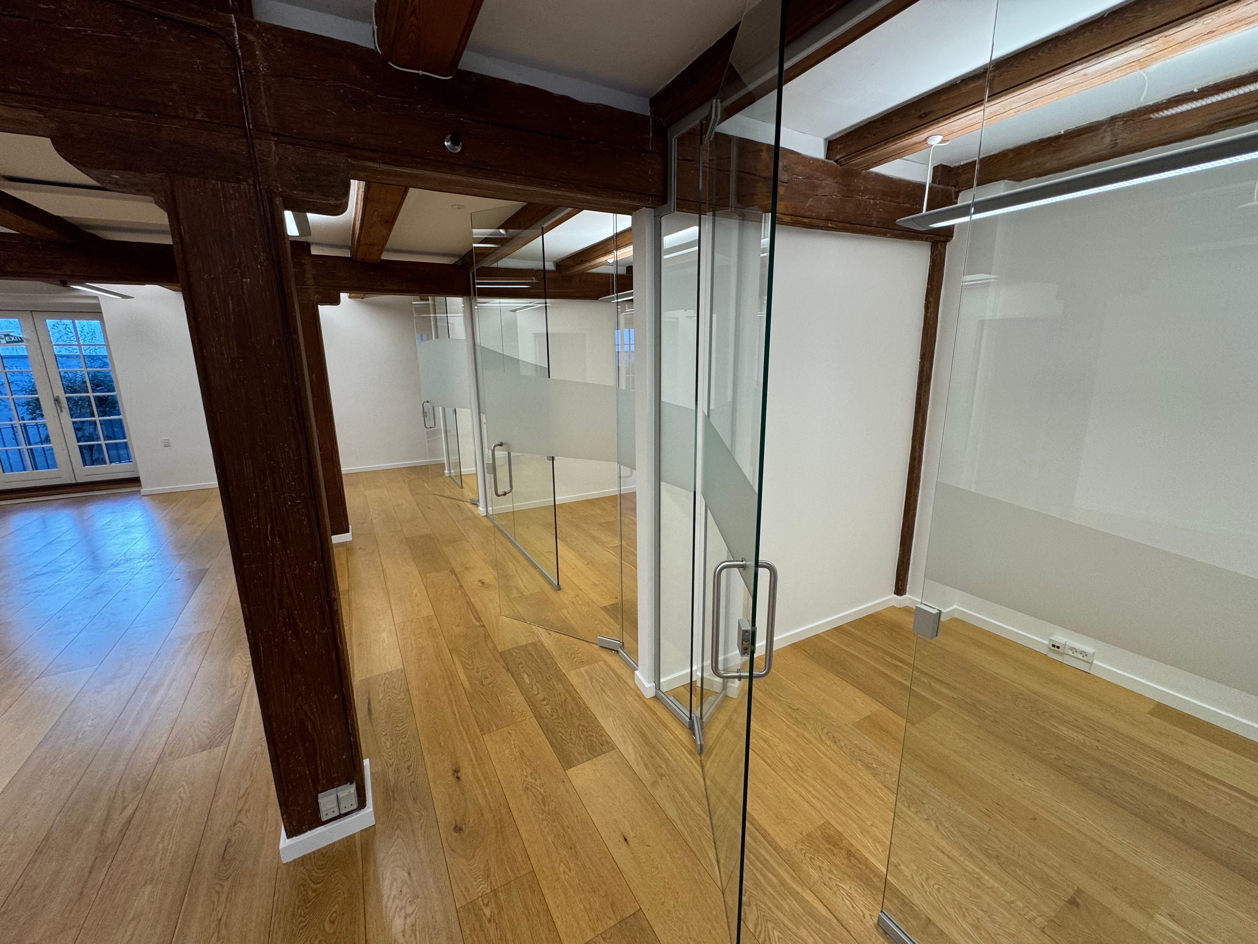 Kontor med lyse loftsbjælke, glas og trægulv samt hvide vægge og hvide døre. Ingen møbler og alt er tomt.