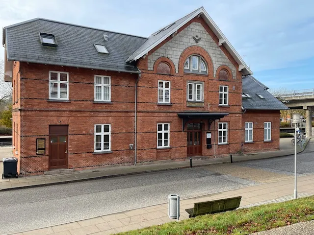Togstation i røde mursten med flere vinduer og en vej foran. To skraldespande og en bænk uden for.