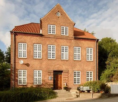 Et stort, rødt murstenshus med flere store vinduer og et rødt tag med en indgang dækket af træ og en plante til højre