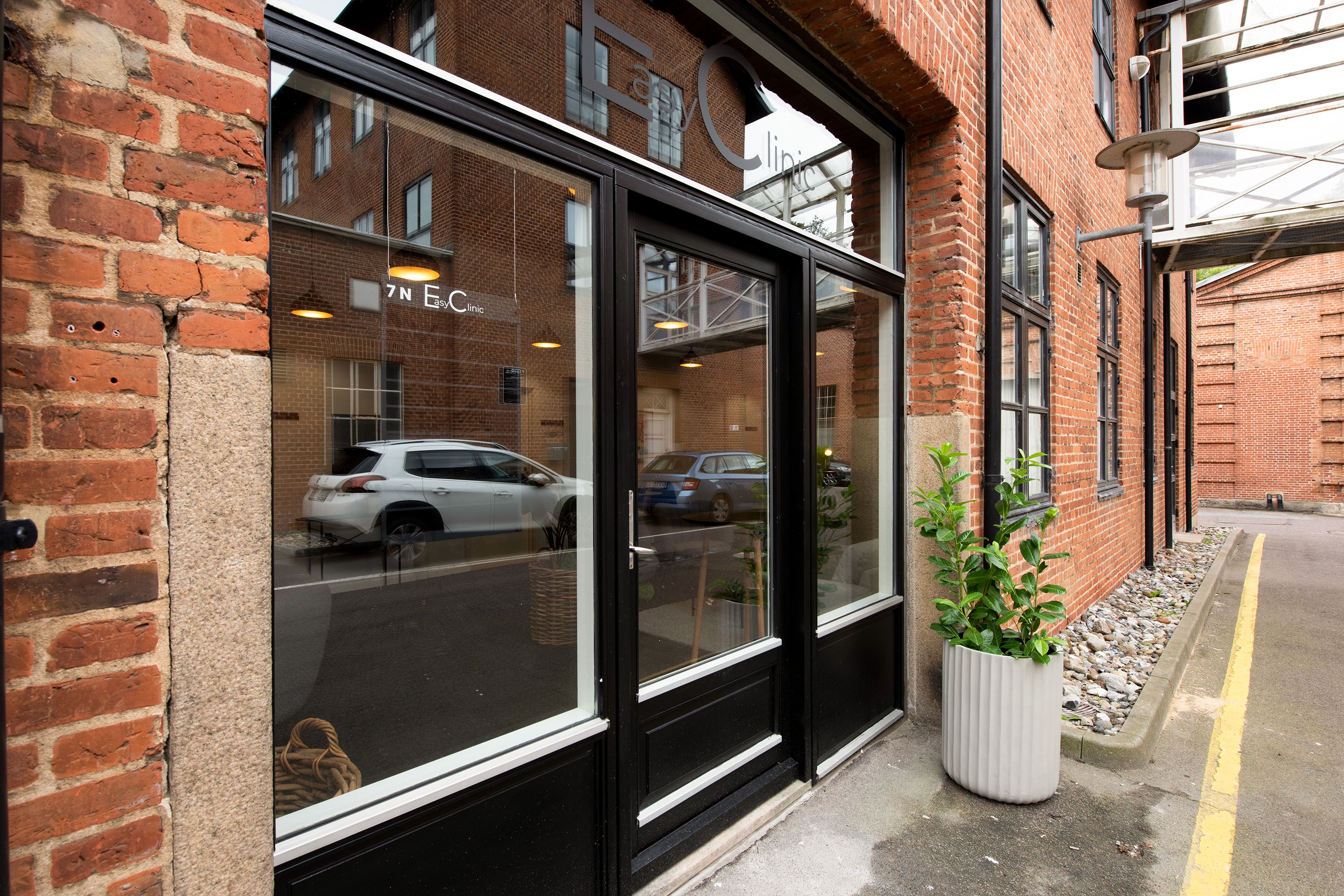 En glasdør med en refleksion af en bil og et hvidt potteplante udenfor en bygning med røde mursten og teksten "Easy Clinic"
