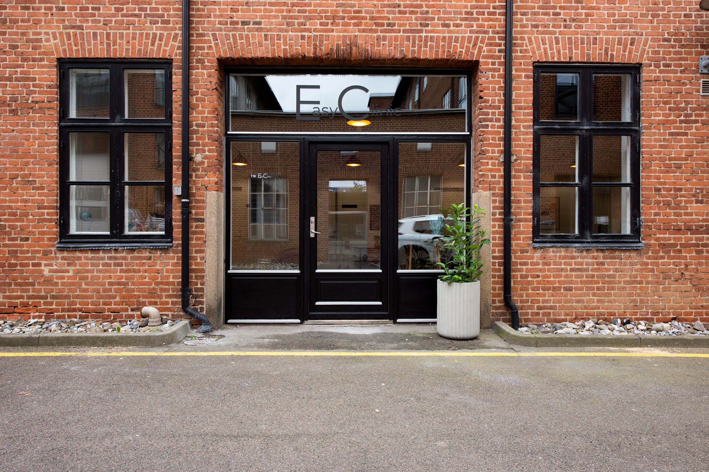 En rødmaursten bygning med store vinduer og en indgang med mærket EasyClinic og en plante i en hvid potte
