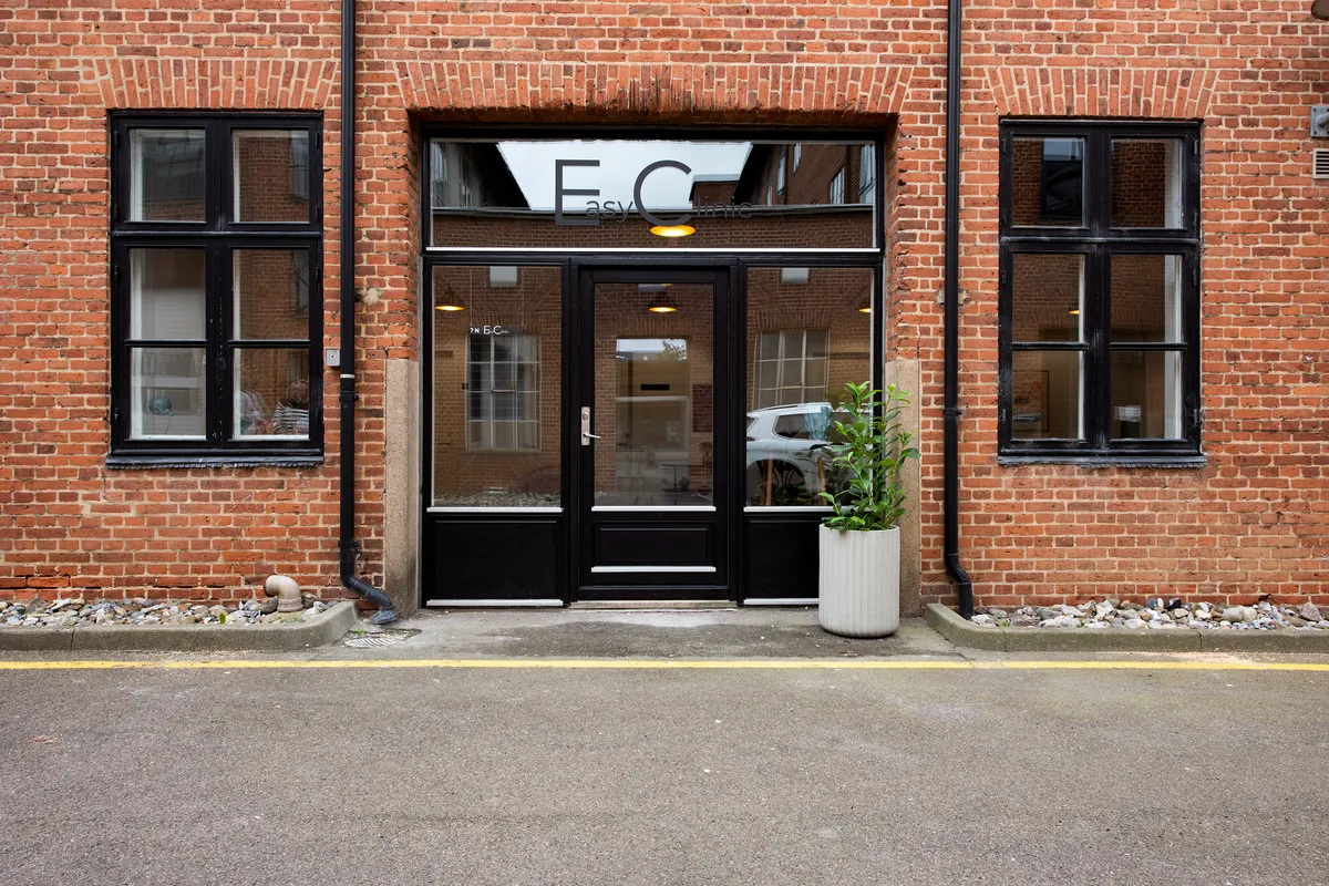 En rødmaursten bygning med store vinduer og en indgang med mærket EasyClinic og en plante i en hvid potte