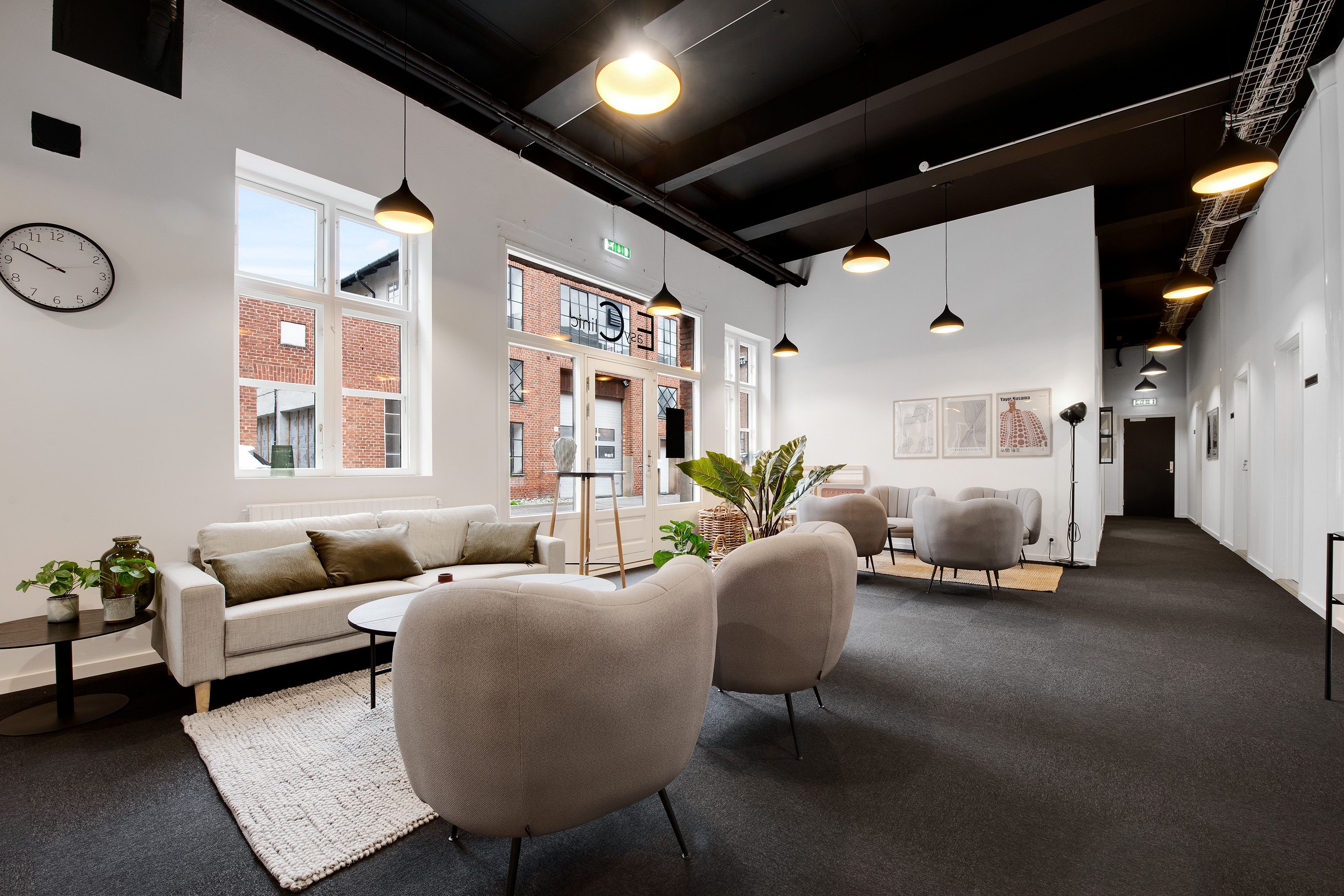 Nydelig lobby med sofaer, plante og lysekroner i lysegrå farve. Væggen er hvid og der er en klokkeskilt på den. Loftet er
