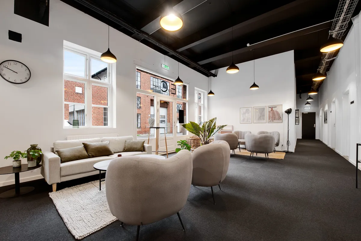 Nydelig lobby med sofaer, plante og lysekroner i lysegrå farve. Væggen er hvid og der er en klokkeskilt på den. Loftet er