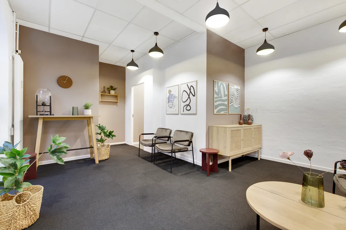 En lille, indbydende venteværelse med to stole, en høj, træskammel, to små runde stole, og en rund, høj træbord med blomster i en grøn vase