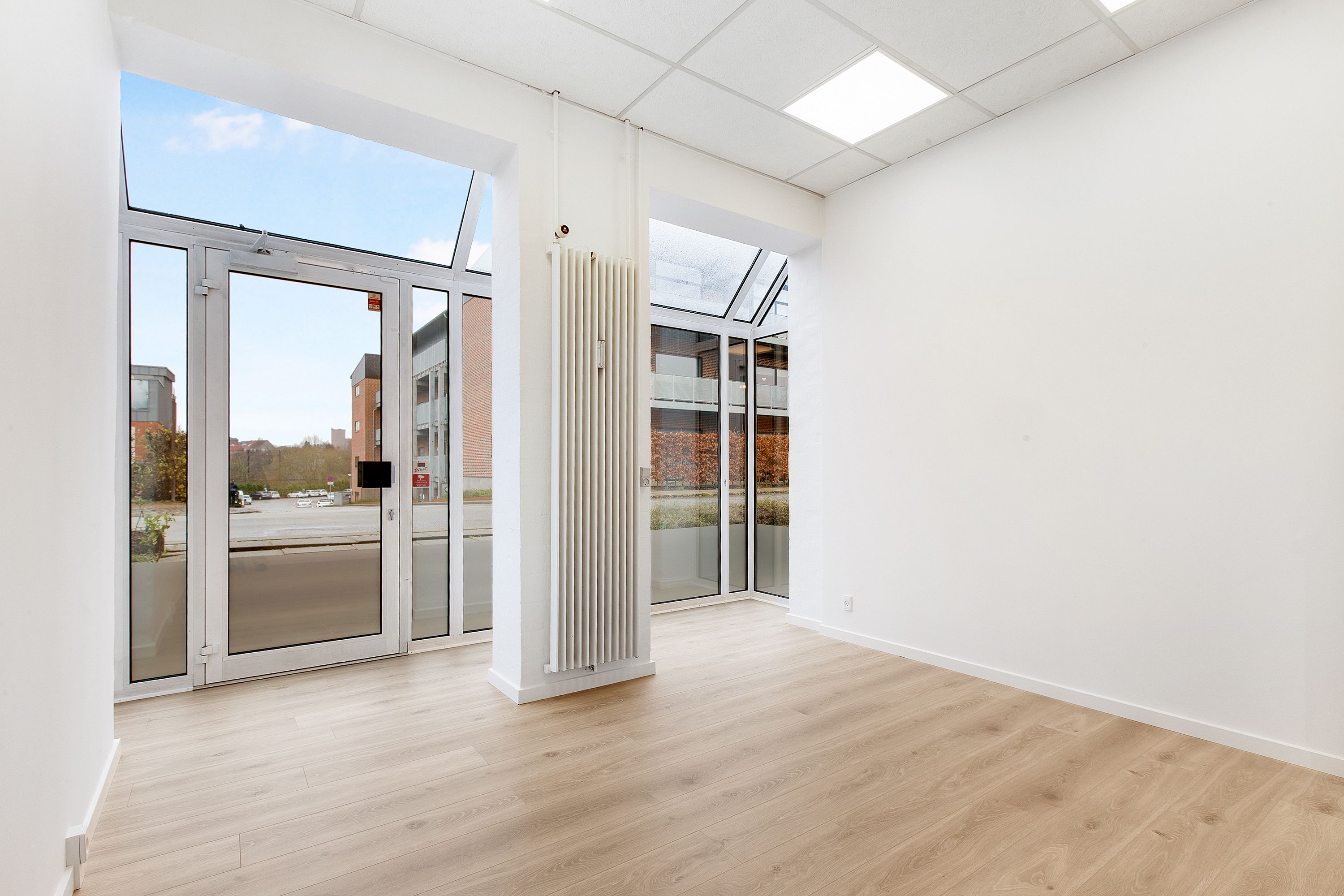 Ledig rum med lyseruder og et stort glasdækket vinduesparti med glasdør. Hvid gulvbelægning og hvide vægge med hvid gulvbeskæring. Et hvidt radiator er monteret på væggen