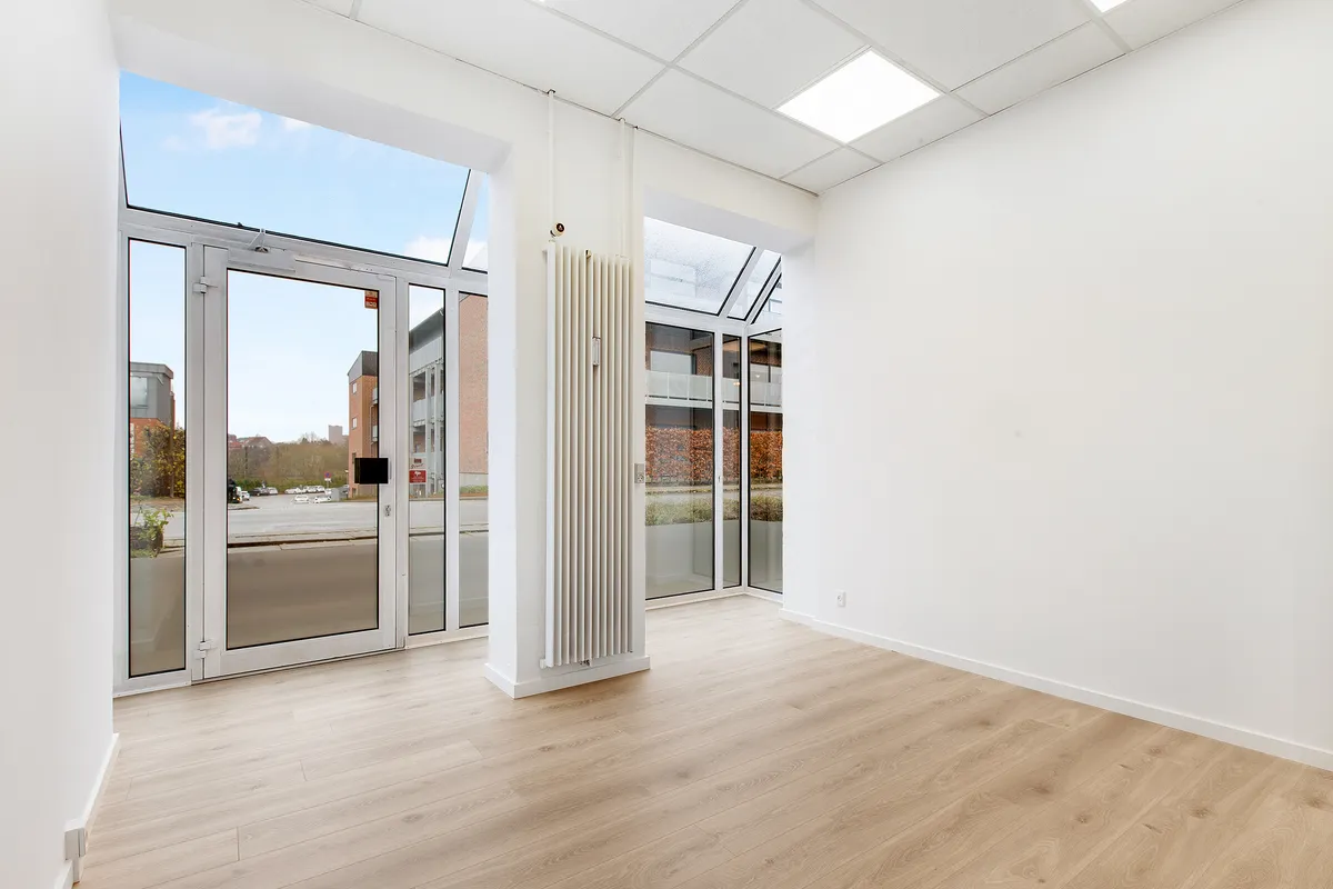 Ledig rum med lyseruder og et stort glasdækket vinduesparti med glasdør. Hvid gulvbelægning og hvide vægge med hvid gulvbeskæring. Et hvidt radiator er monteret på væggen
