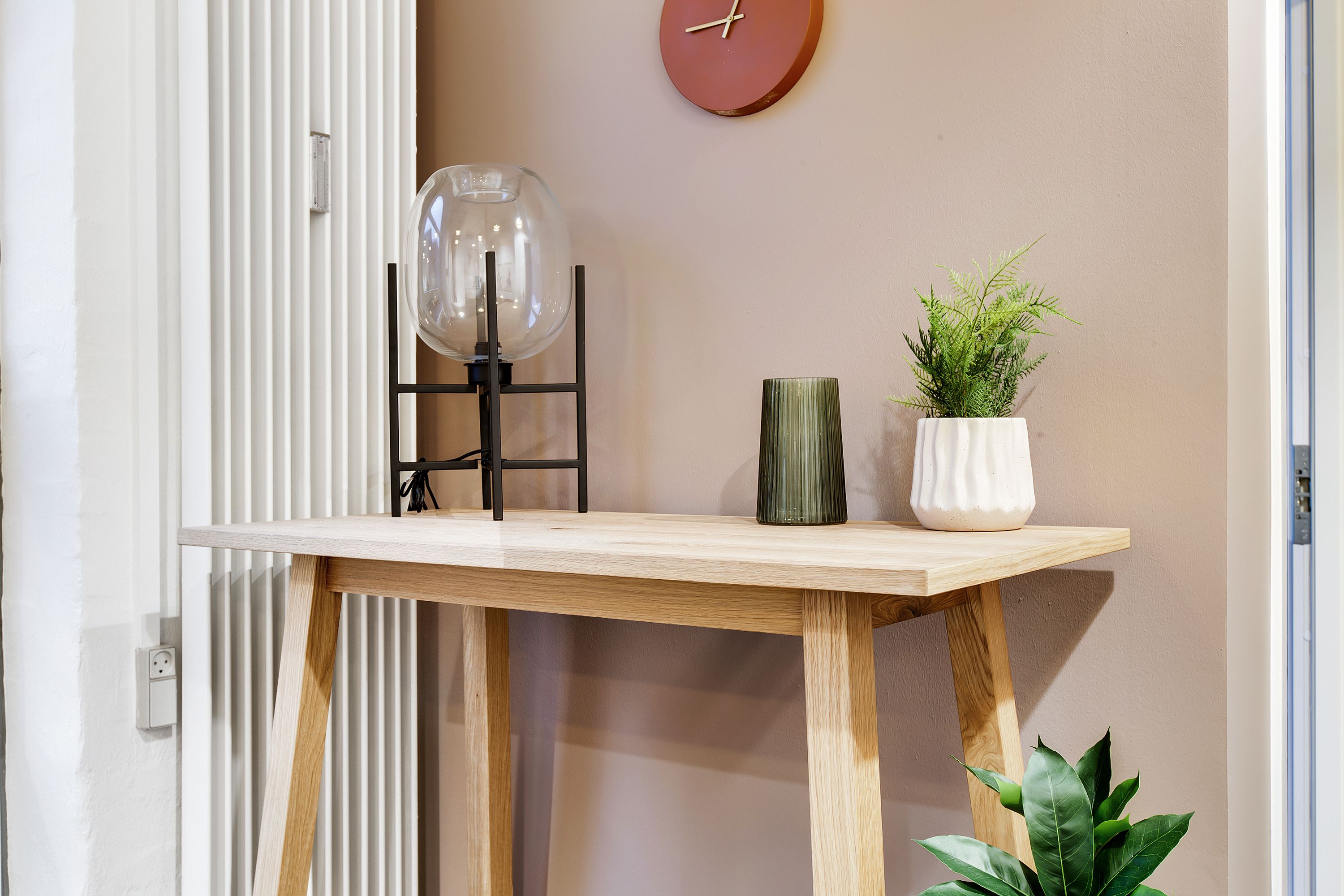 Lejlighed med højloftede rum, elegant konsolbord og plante. Glasskærmslampe og grønt plante i hvid vase. Væg med rund ur og belysning