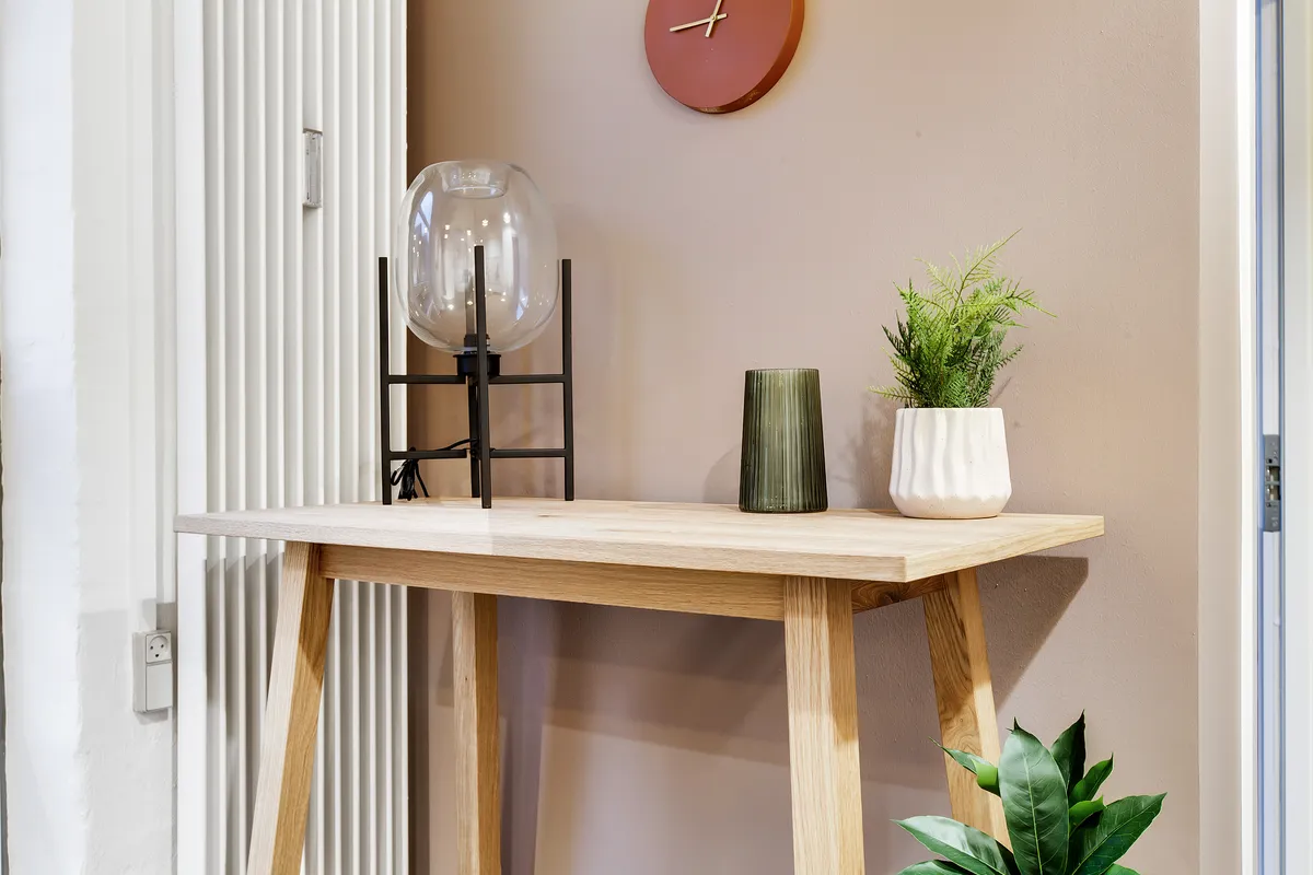 Lejlighed med højloftede rum, elegant konsolbord og plante. Glasskærmslampe og grønt plante i hvid vase. Væg med rund ur og belysning