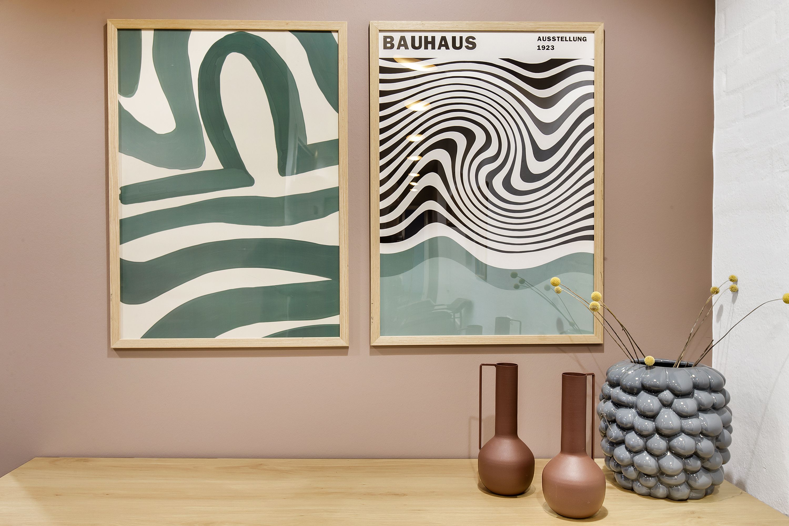 To malerier i Bauhaus-stil er hængt på en lyserød væg. Et maleri har grønne bånd og det andet har sort og hvidt stribet mønster. To vaser med håndtag og en vase med perler er placeret på et bord