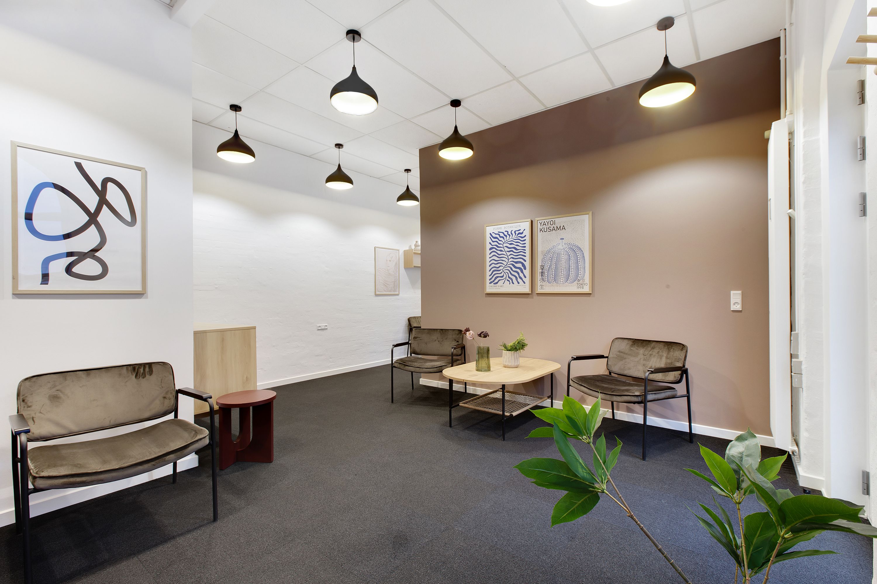 Et rum med lyse vægge, flere møbler og lysekroner samt tre malerier. To store plantepoter med grønne planter og en rund bord med blomster og en vase.