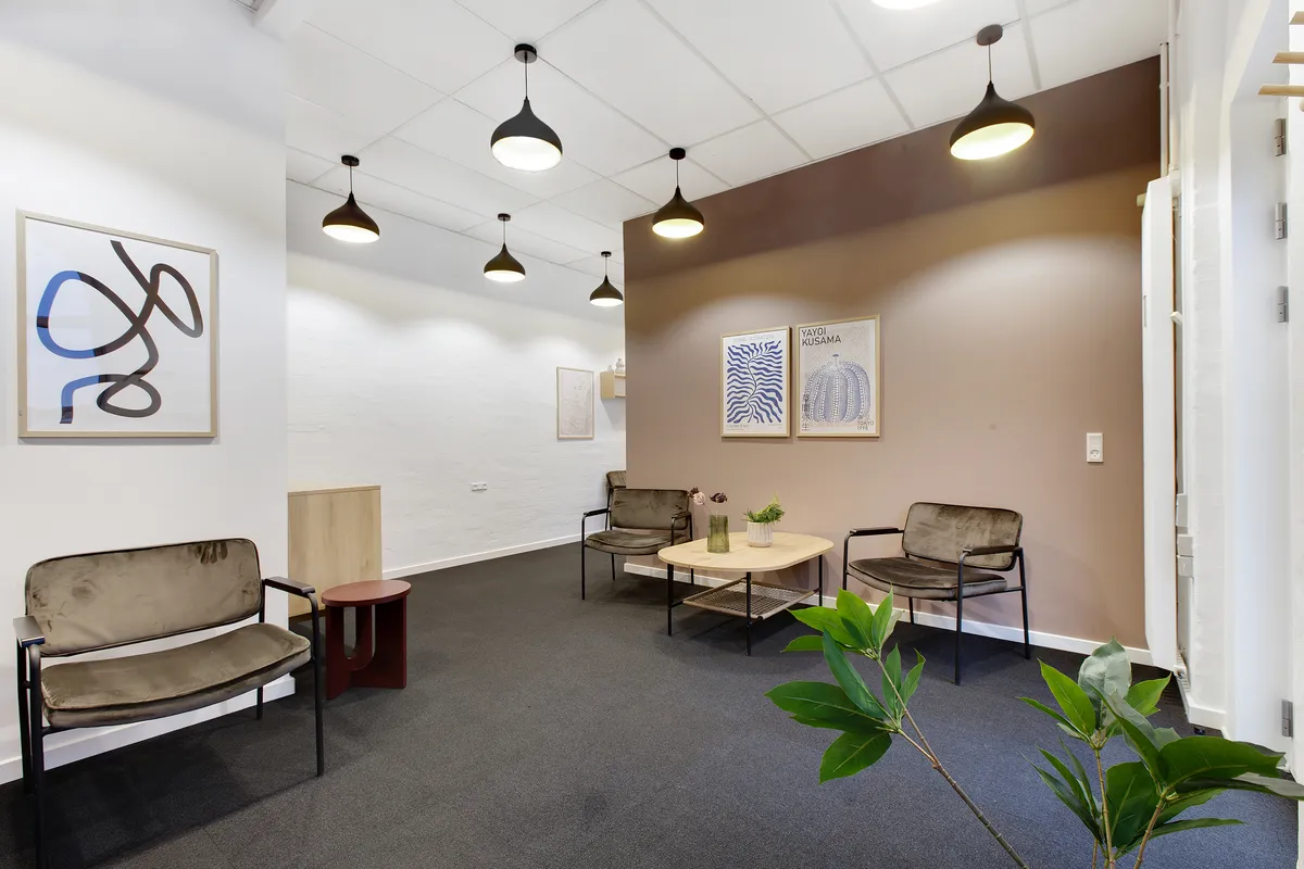 Et rum med lyse vægge, flere møbler og lysekroner samt tre malerier. To store plantepoter med grønne planter og en rund bord med blomster og en vase.