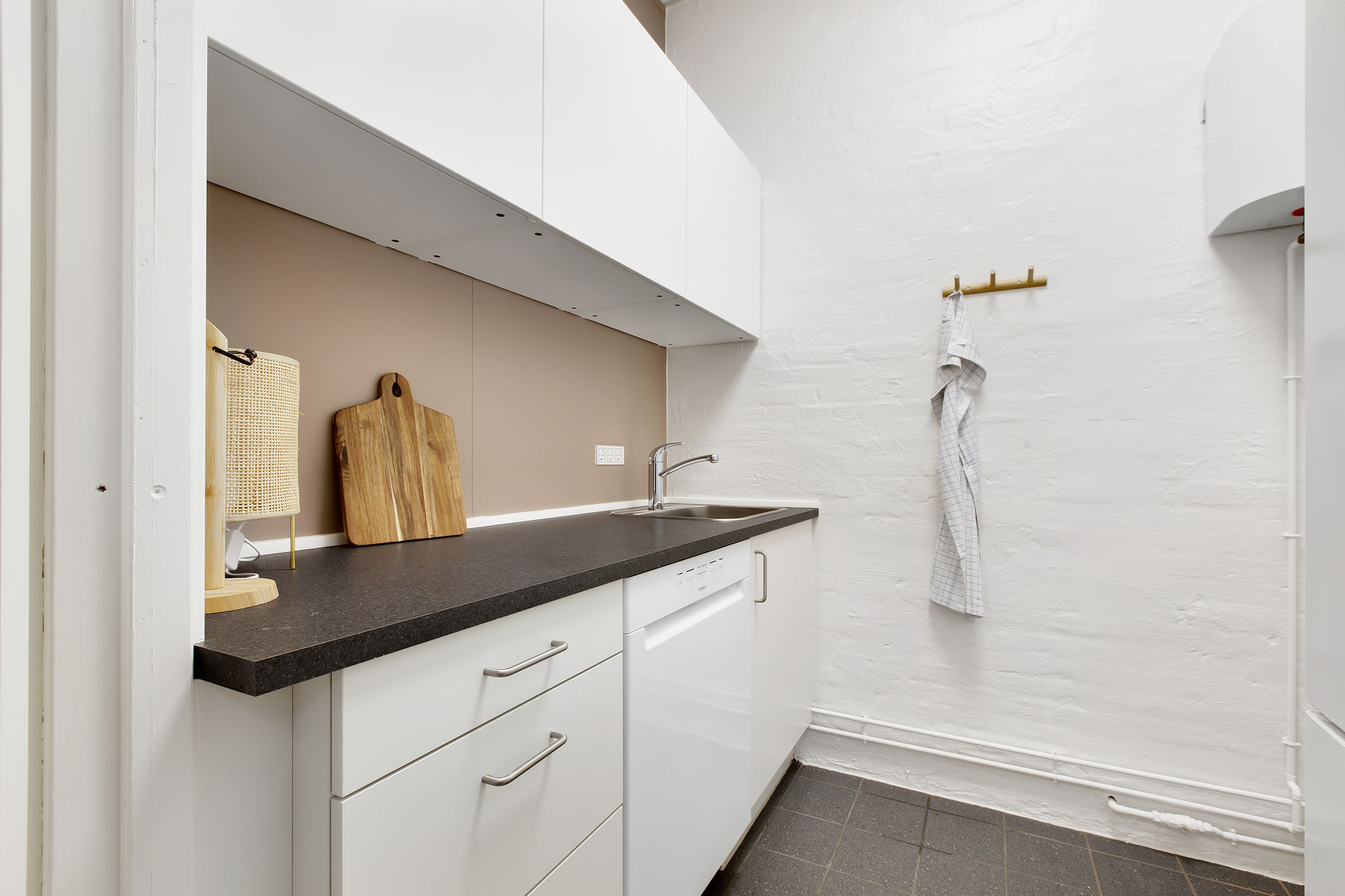 Moderne køkken med hvide vægge og loft, lysegrå gulvfliser, en hængende hvid skuffe, en stående træskærer og en hvid ovn på væggen