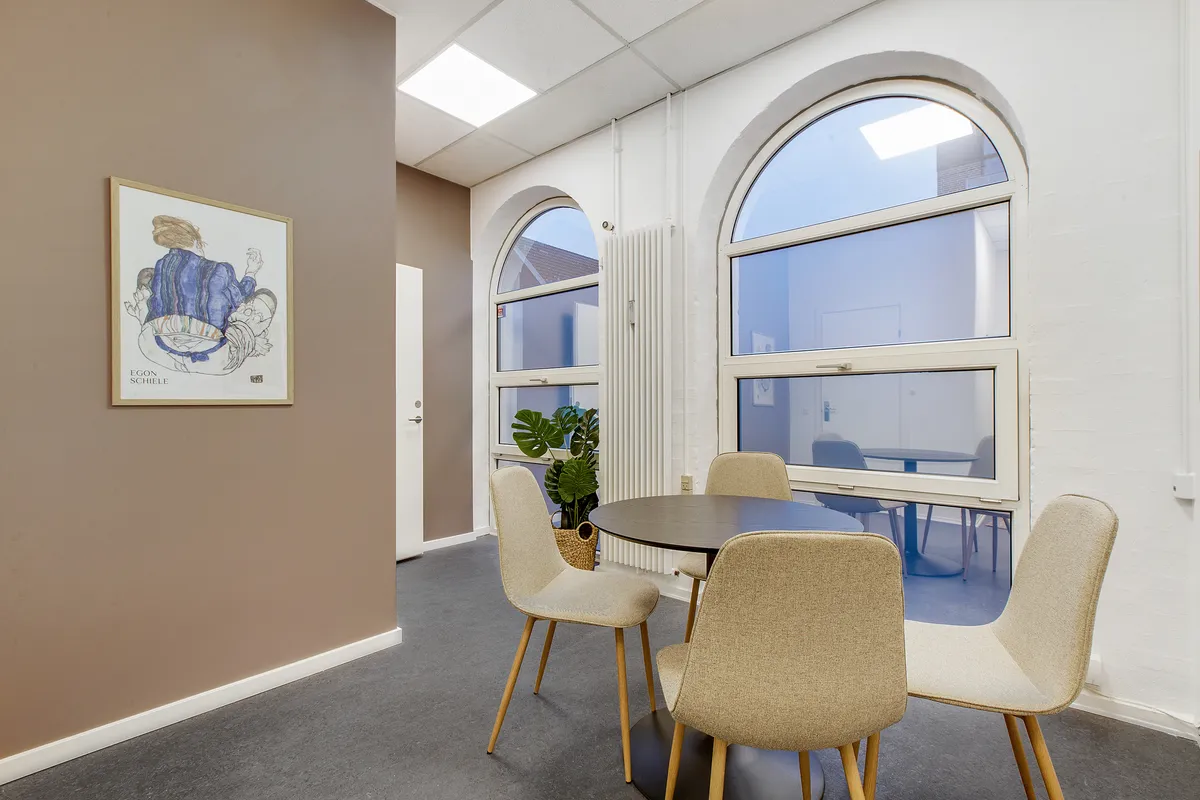 En kontorfællesskab med tre stole omkring et rundt bord, plante i kurv, et maleri og en glasdør med udsyn til et lysere rum