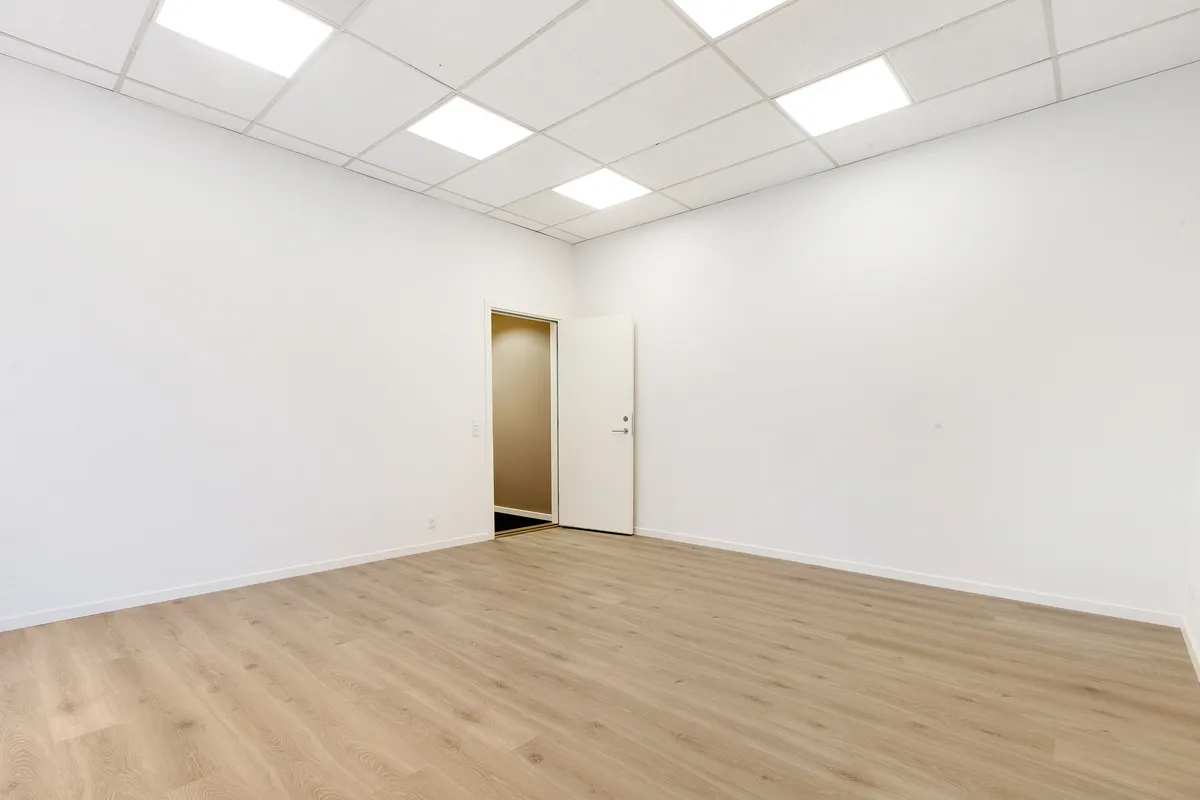 Ledig og fuldt renoveret kontorplads med højloft og trægulv. Åbent rum med hvide vægge og lyskryds. Der er en dør i højre side og en i baggrunden.