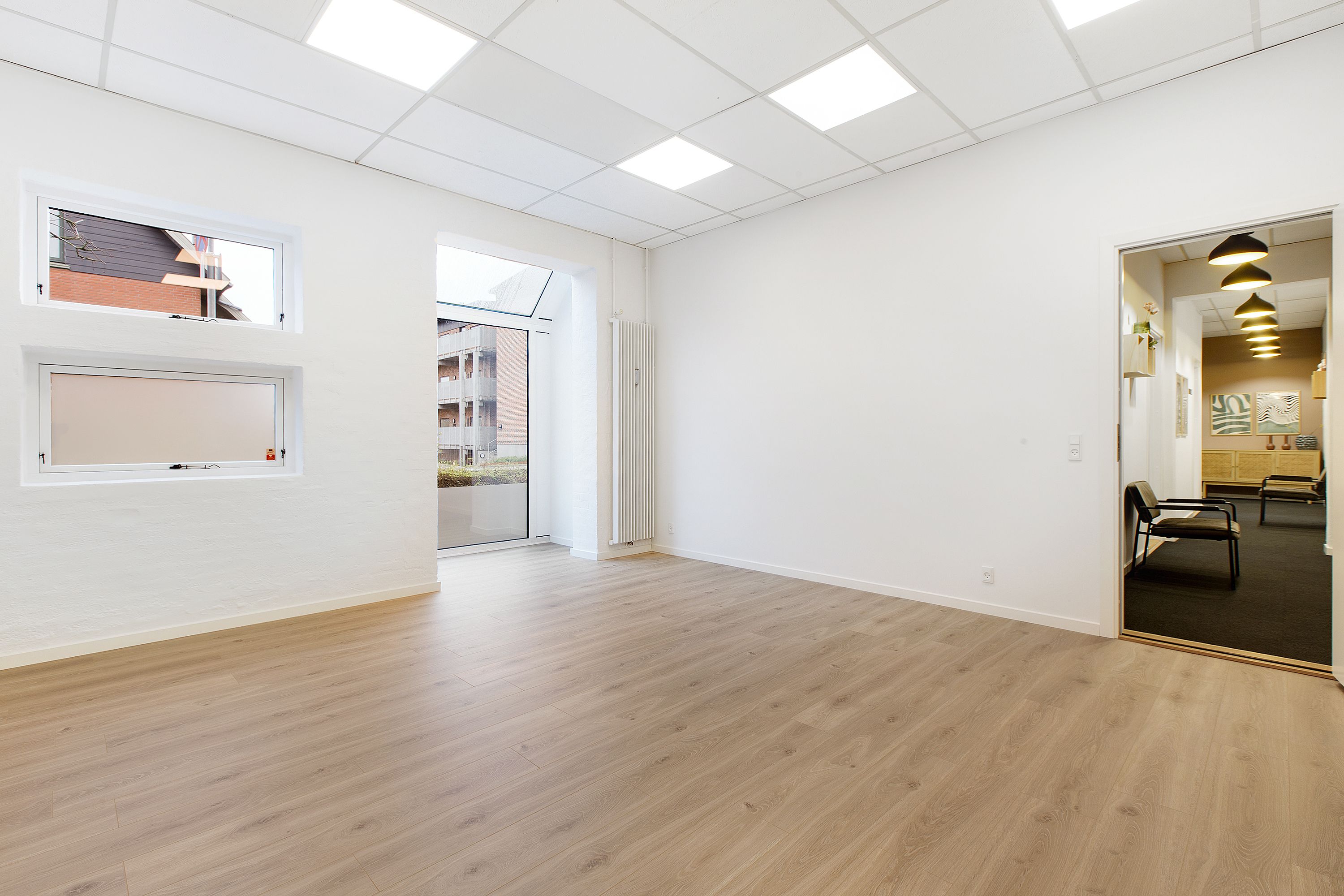 Klinkfællesskab i Kolding med to vinduer, åben dør, hvidt loft, hvid væg, trægulv og moderne interiør med hængende lamper og en lille sofa