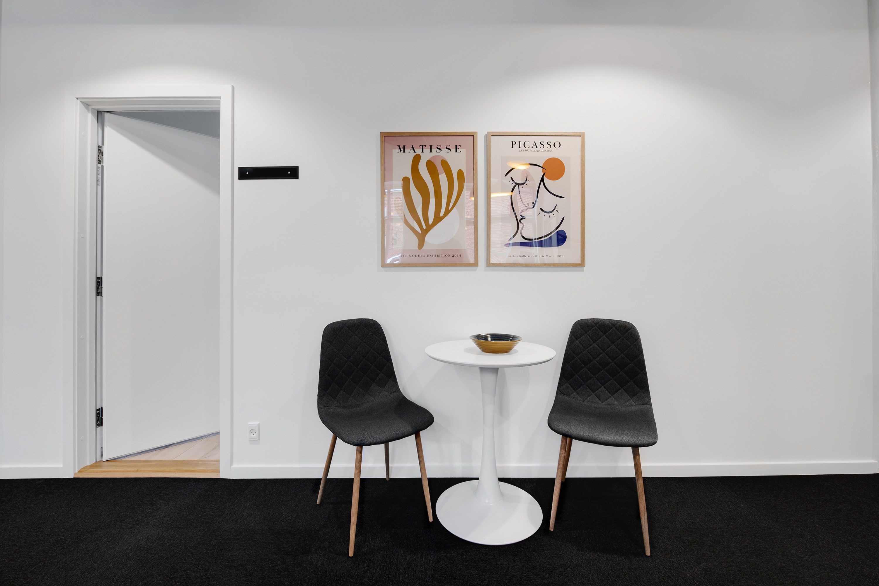 To stole med pudsede sæder og et rundt bord med en skål i midten og to malerier med Matisse og Picasso på væggen