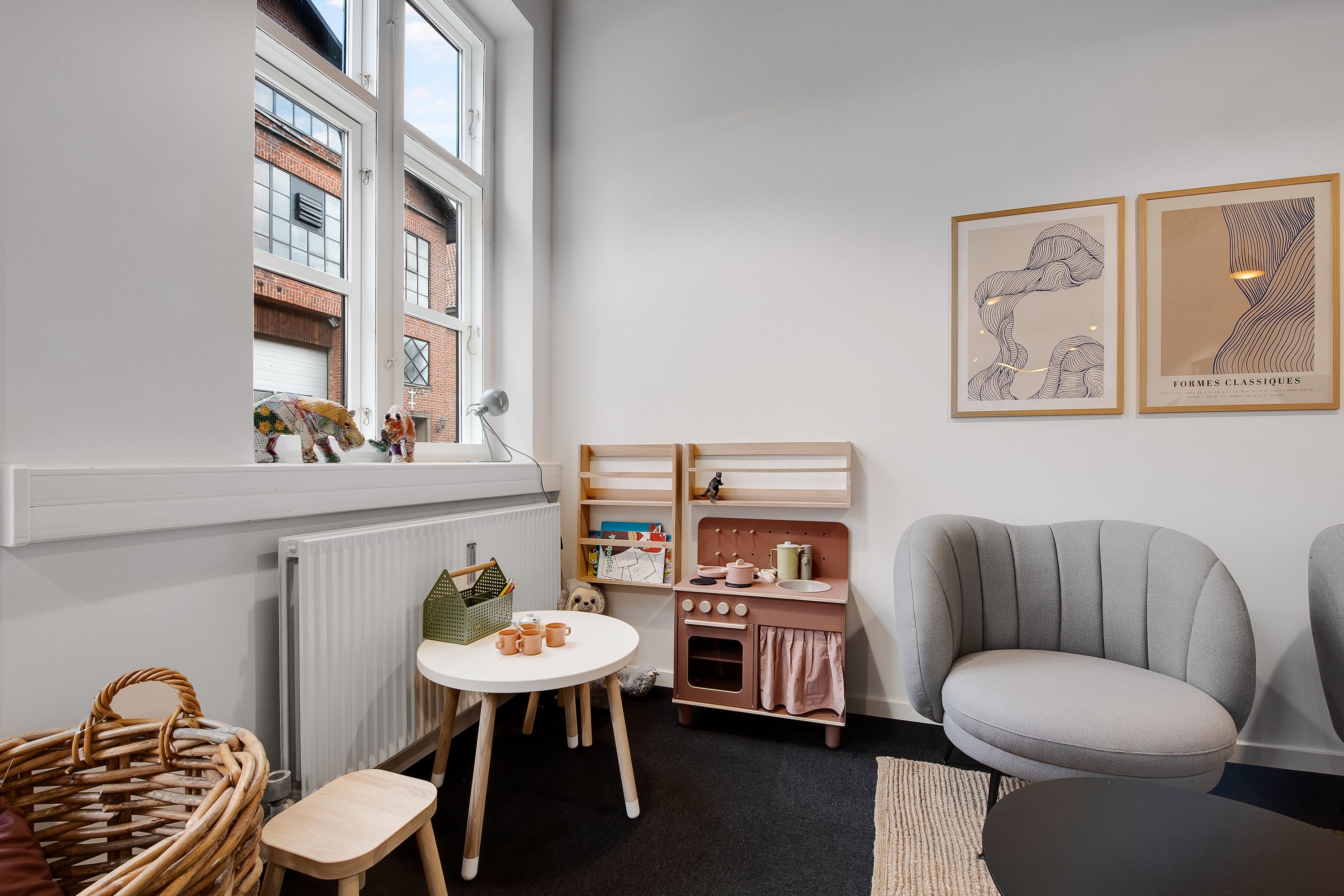 En trækøkken med hylder og bønner, der står ved en hvid væg med malerier og en radiator samt en rund hvid bord og en grå stol