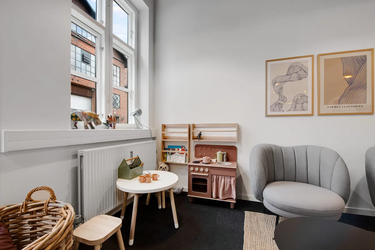 En trækøkken med hylder og bønner, der står ved en hvid væg med malerier og en radiator samt en rund hvid bord og en grå stol