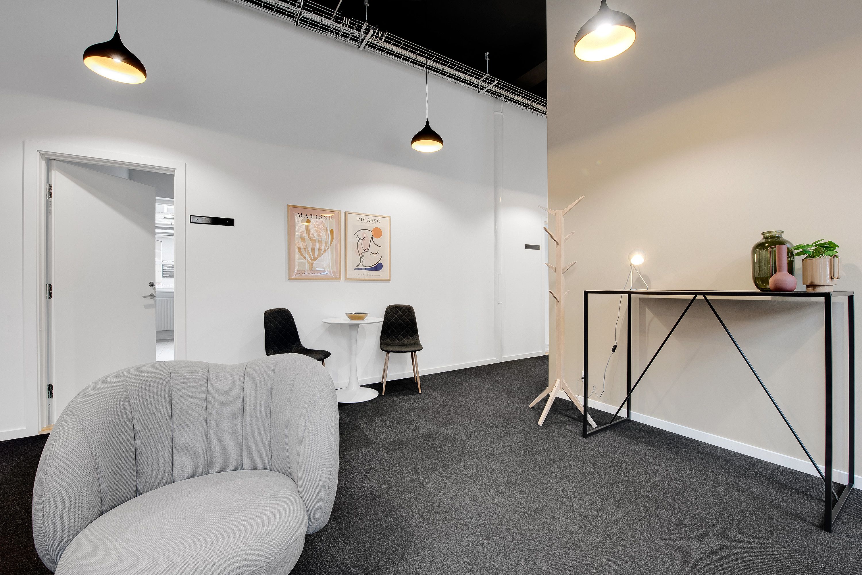 En moderne kontorrum med en hvid væg, en hvid dør, og et lysekroner hængende fra loftet. Et højt bord med to glas og en vase