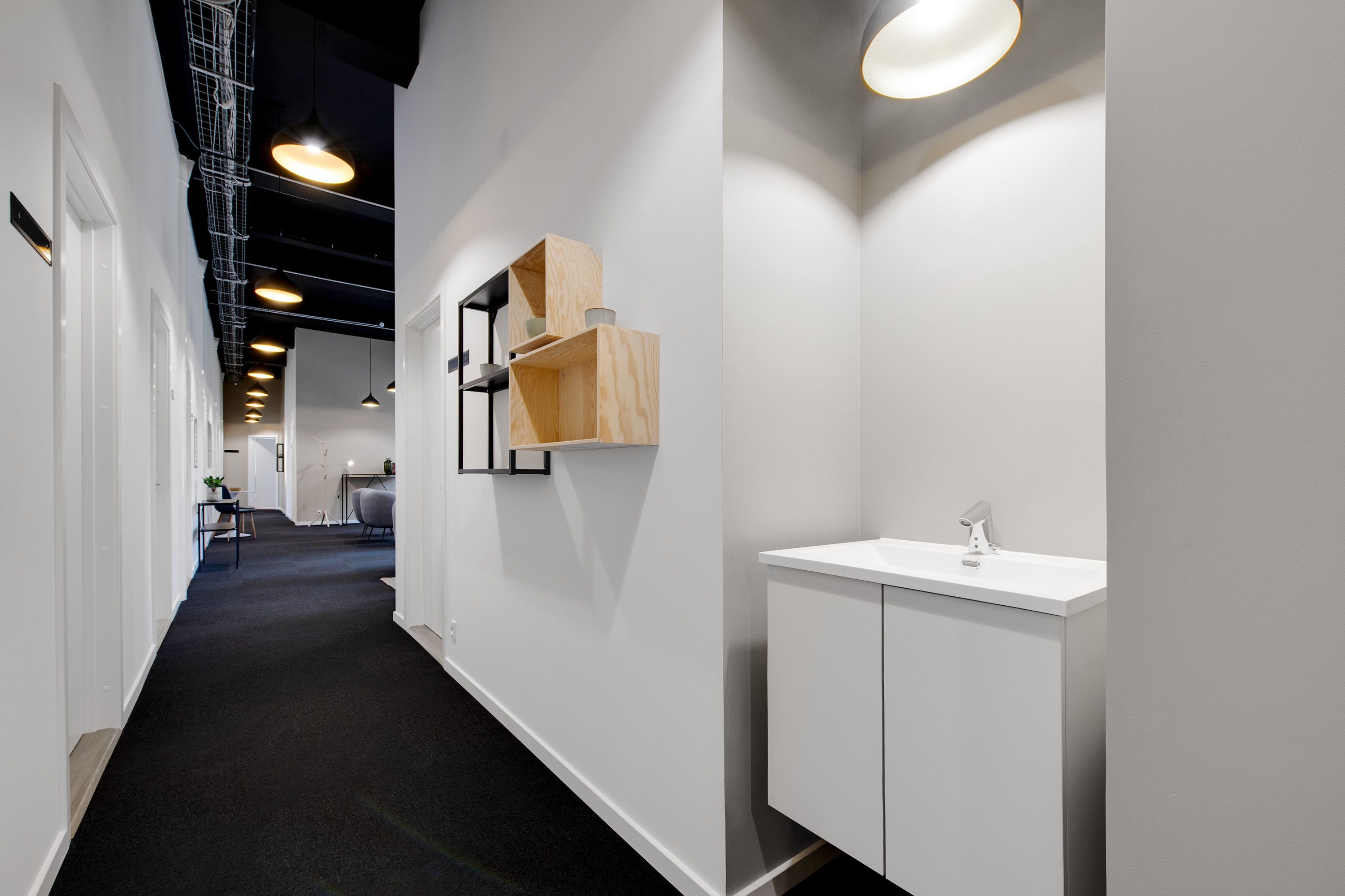 Et smal korridor med hvide vægge, gulv og loft, og møbler i minimalistisk stil, samt et toilet med hvid vask og lysekroner