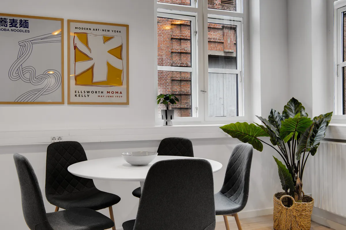 Et moderne og minimalistisk spisestue med rundt bord og fire stole omkring. Der er to plakater på væggen, og der er en stor