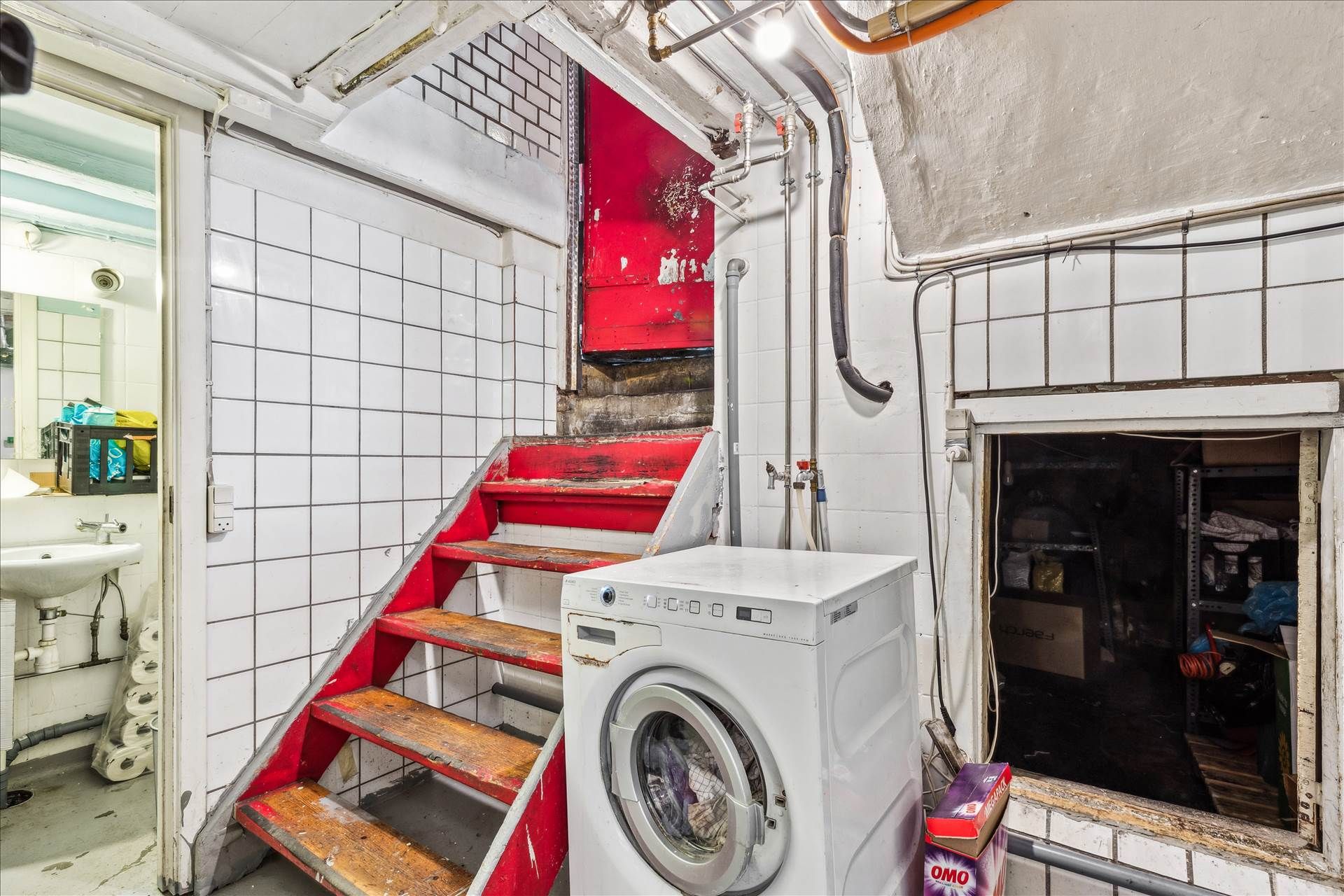 Vaskemaskine ved siden af rødfarvede trapper, der fører ned til kælderen med åbne døre og indvendige genstande