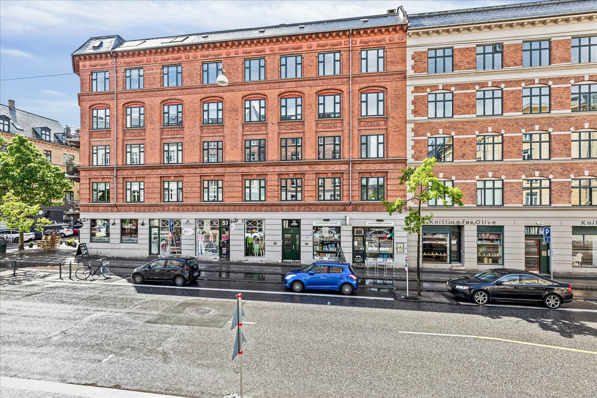 Biler parkeret foran bygning, tekstforretning "Knitting for Olive" og reklameskilt "Candido" med cafeindgang, gågade med