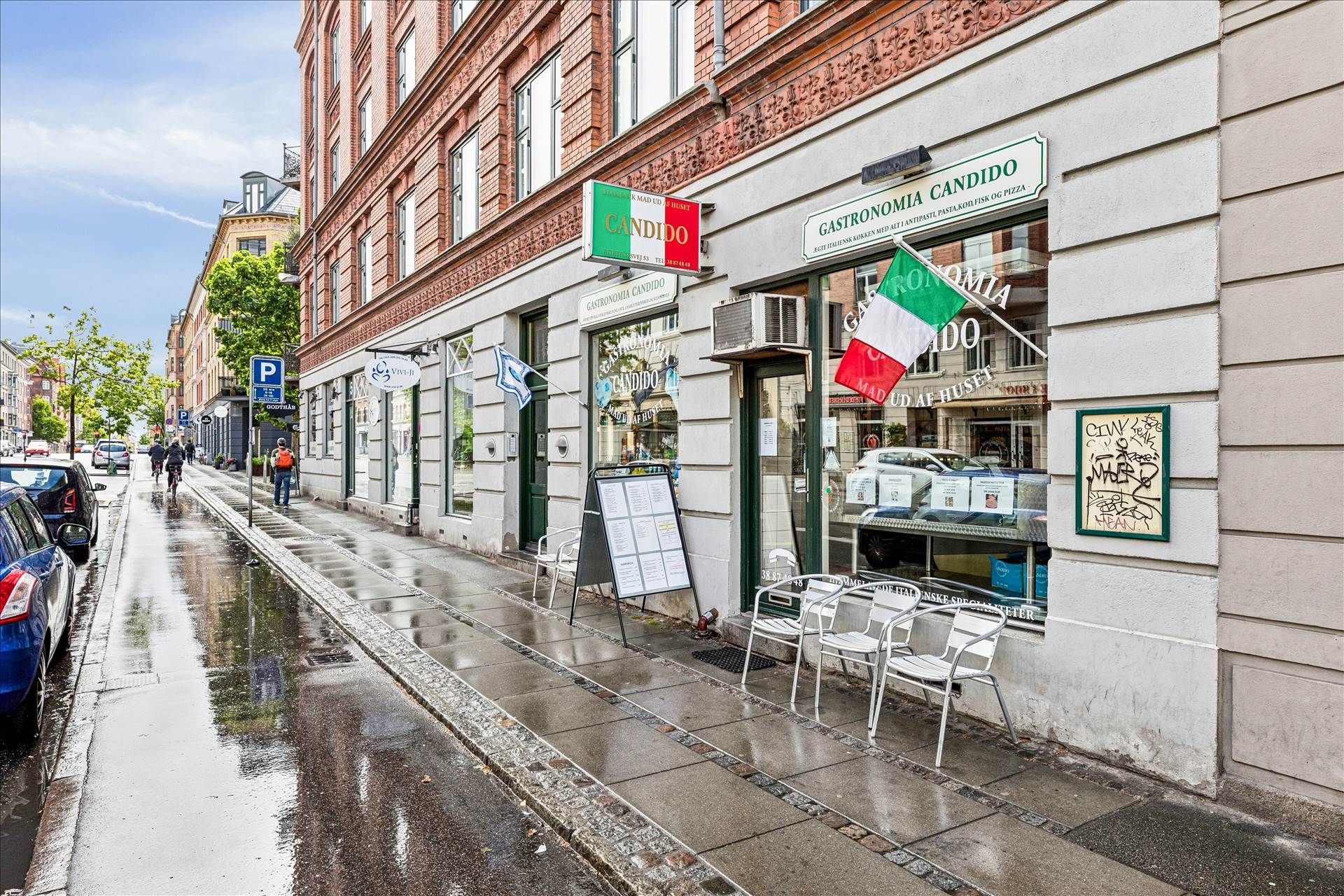 En italiensk restaurant med navnet Candido, hvor der er flag udenfor, og der er parkerede biler og gående mennesker på
