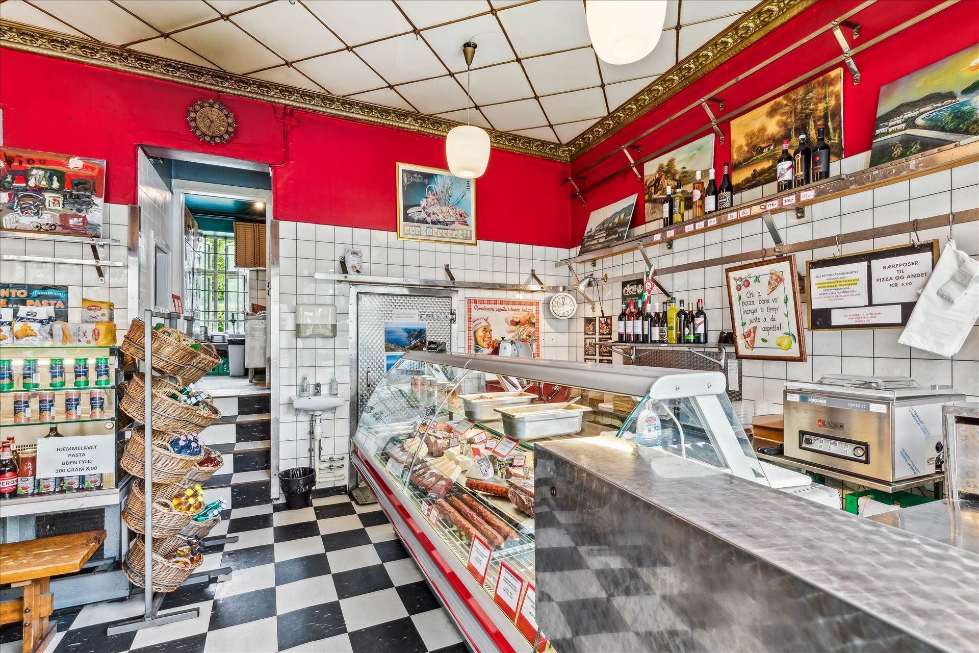 Køkken med sort-hvid tæppegulv og røde vægge, delikatesseudstilling med oliebøtter og pølser, pastahylderne, og gulvet er