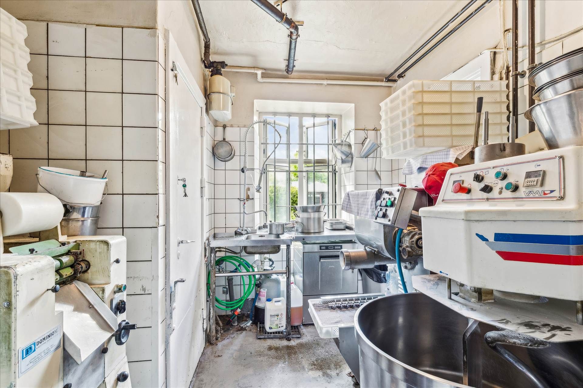 Køkken med flere maskiner og beholdere, og en åben dør med en slange på gulvet og en vindue med metalruder