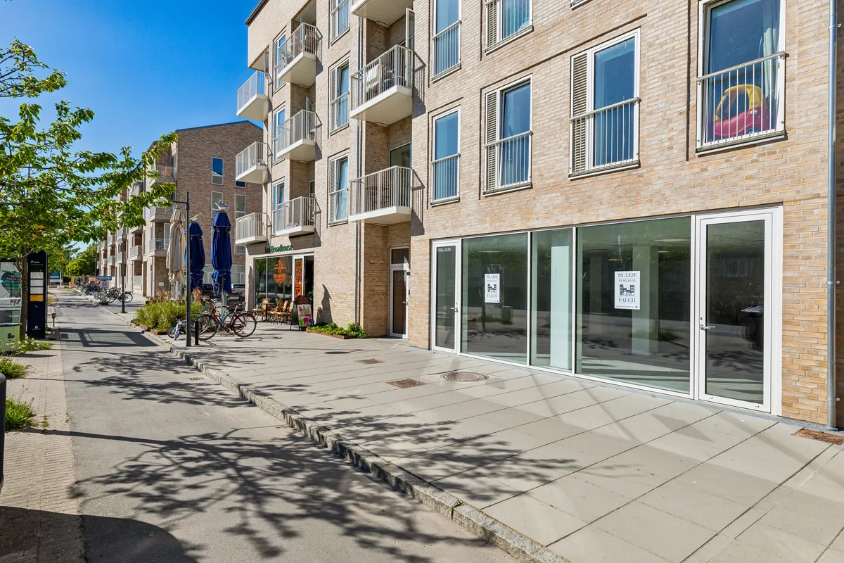 Butikslokaler i et nyt boligområde med balkoner og plantager foran. Gammel og ny bebyggelse langs gågaden med både cykler og