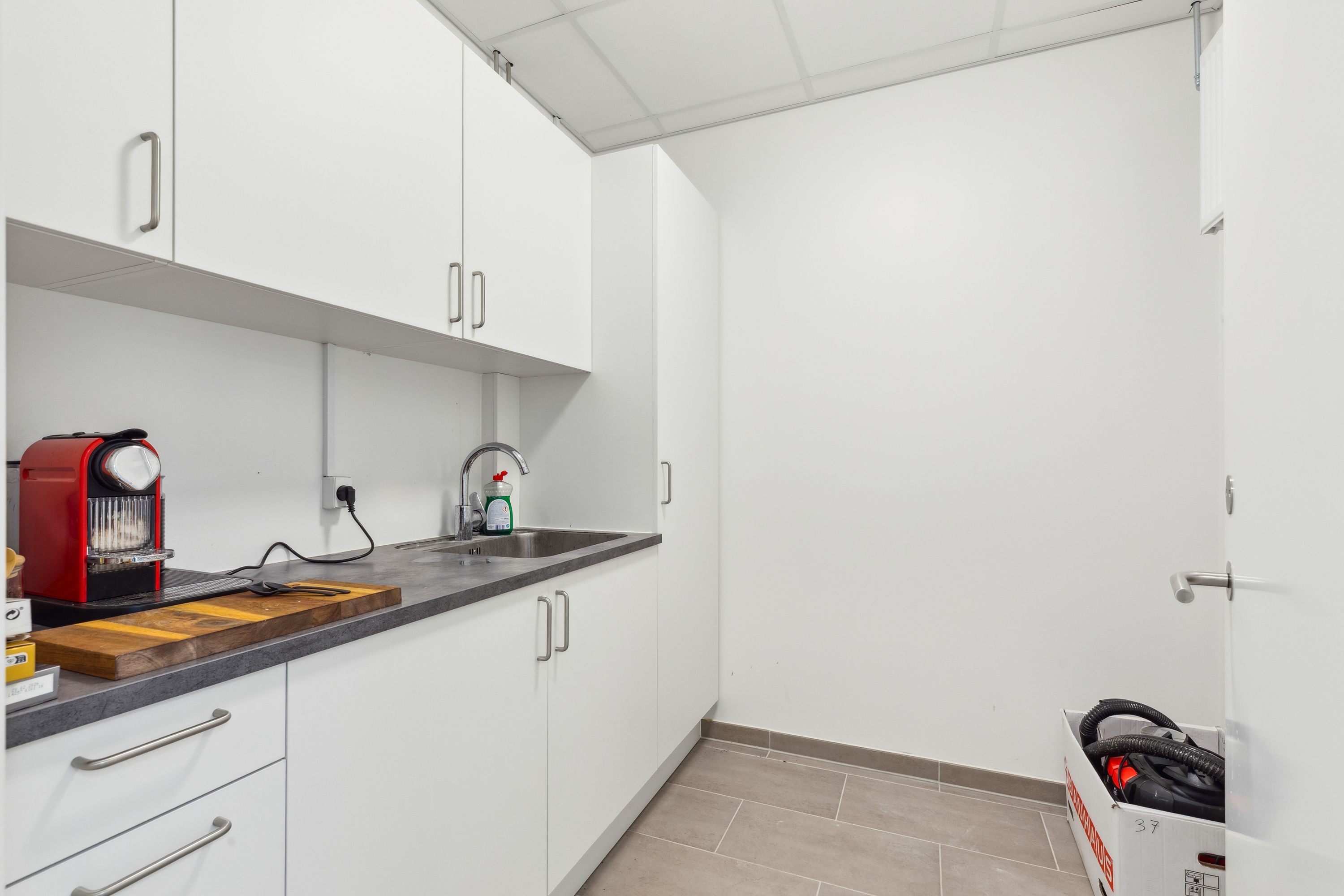 Foto af et lille køkken med en rødt og sort espressomaskine, en træskæreplade, en sæbeflaske og en børste samt håndkle og en