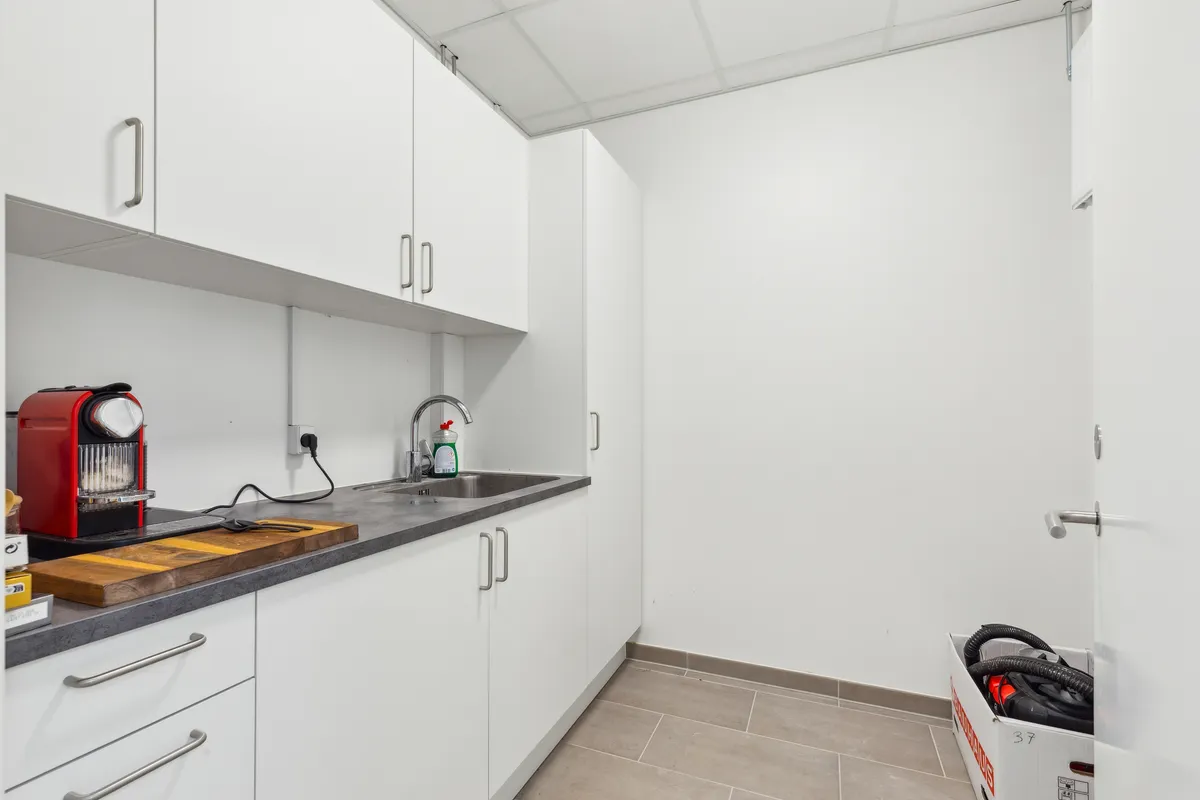 Foto af et lille køkken med en rødt og sort espressomaskine, en træskæreplade, en sæbeflaske og en børste samt håndkle og en