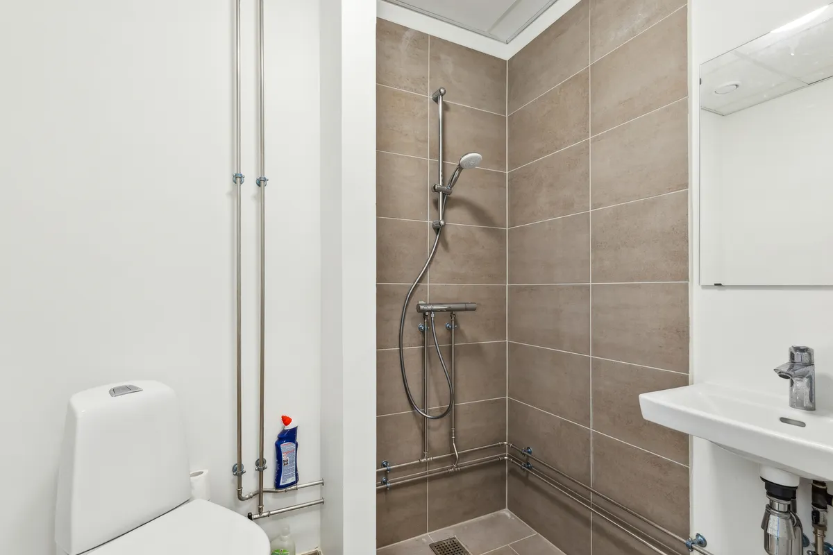 Ren og moderne badeværelse med hvid toilet, hvidt vask og brusebad med dobbelthovedsbruser og en lille flaske med