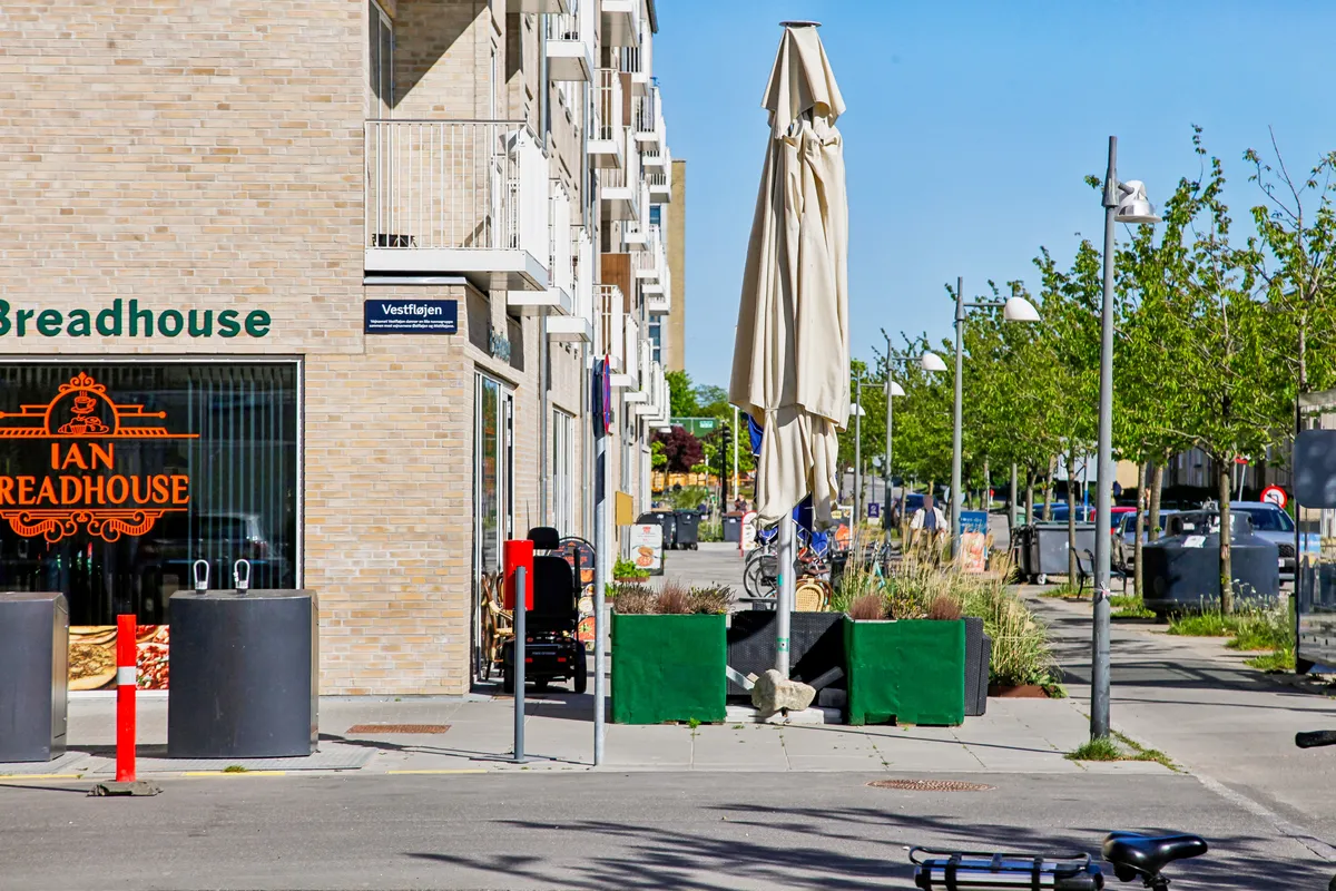 En række huse med facade mod fortovet, foran Breadhouse med en åben paraply og plantepotter, samt en cykelparkeringsplads