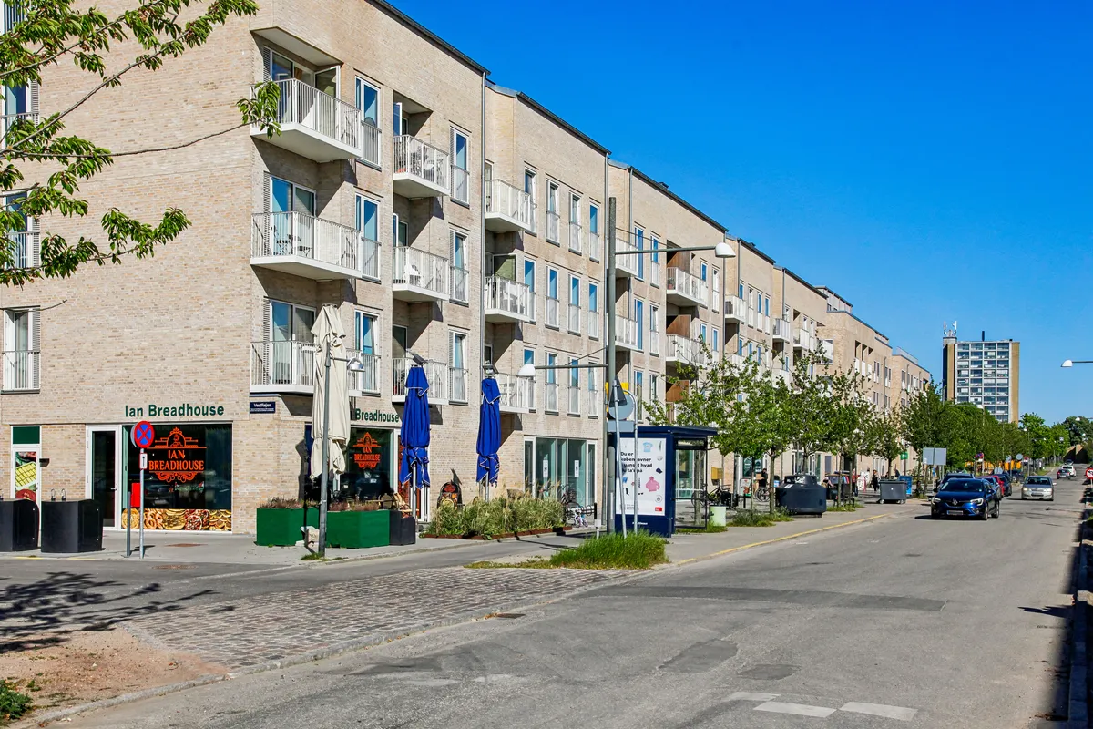 Bred gadegade med moderne lejlighedsbygning med udsigt til Ian Breadhouse bagefter flere biler parkeret og folk går