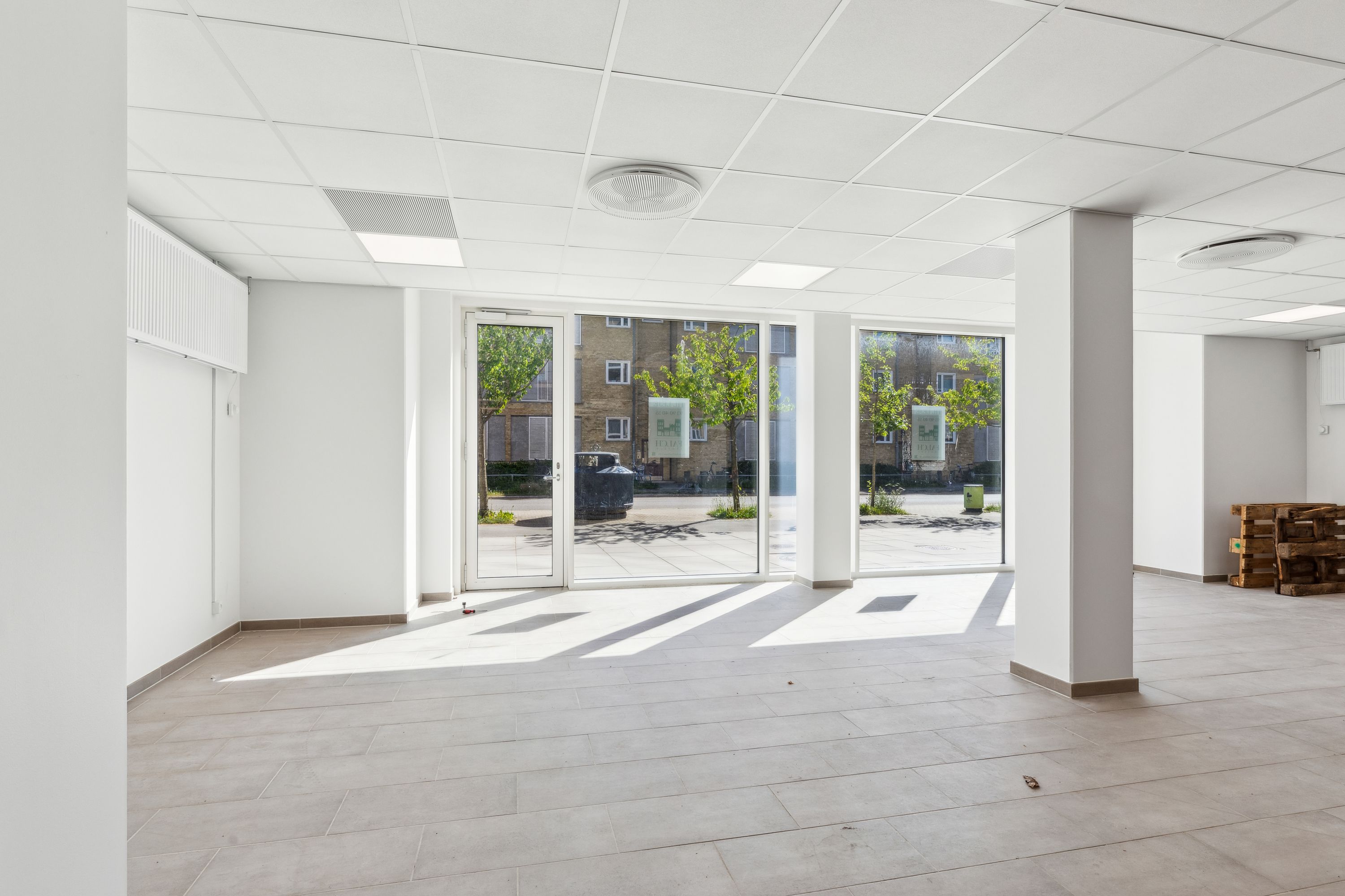 Ledig lokale i ny ejendom med store vinduer og træpalé. Interiør med hvid gulv og vægge samt loft. Styret lys og rekvisitter