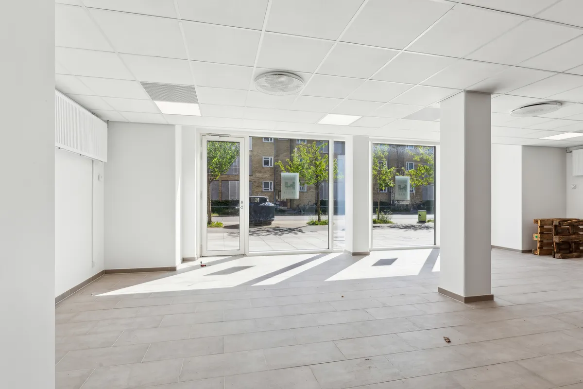 Ledig lokale i ny ejendom med store vinduer og træpalé. Interiør med hvid gulv og vægge samt loft. Styret lys og rekvisitter