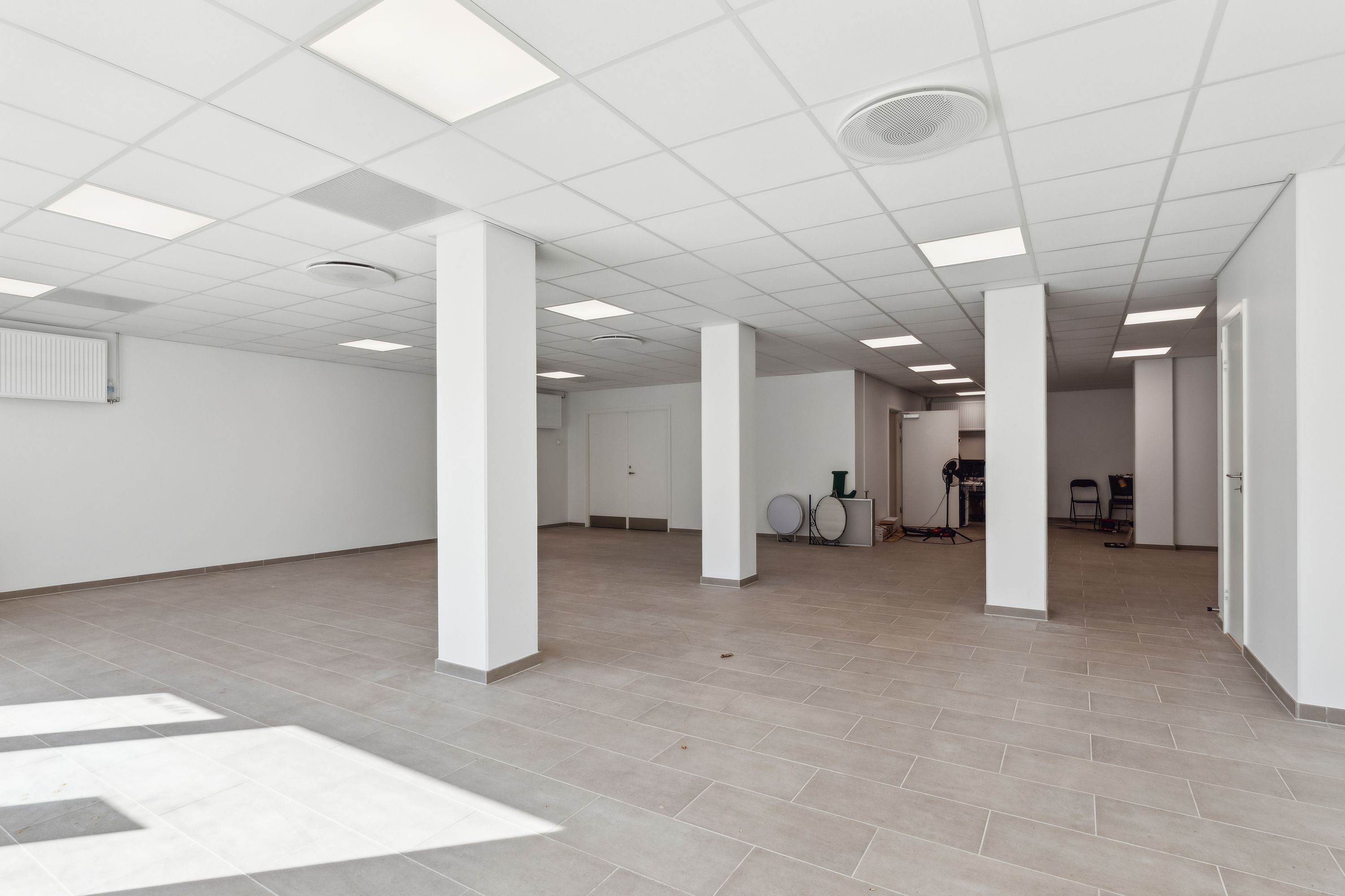 En stor, lys, tom stue med hvidt loft, hvide vægge og gulv med lysegrå fliser. I hjørnet er der en garderobe og en åben dør.