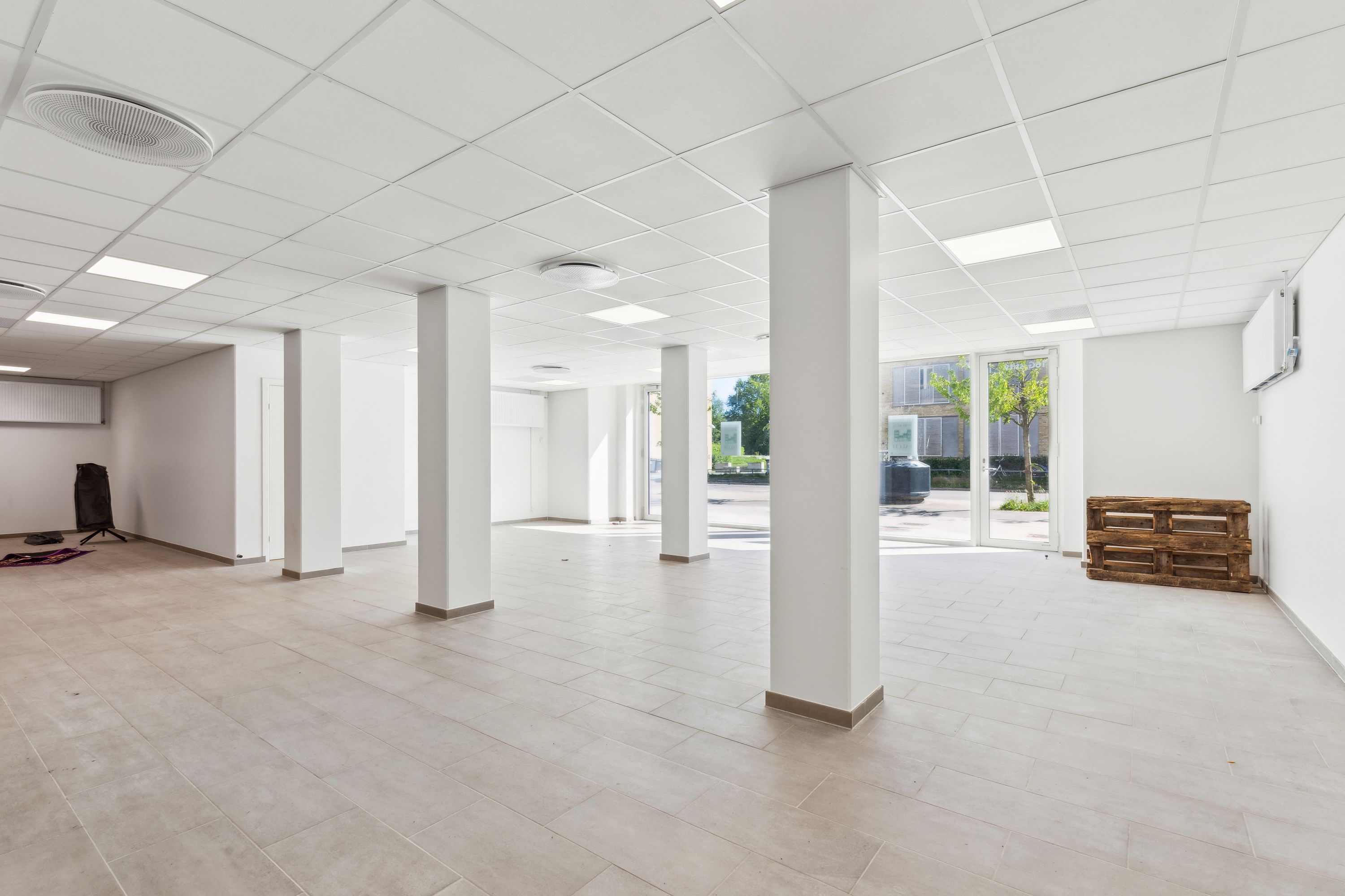 Ledig lokale med højt til loftet og hvide vægge, store glasdøre mod gårdhaven og træpalle i hjørnet